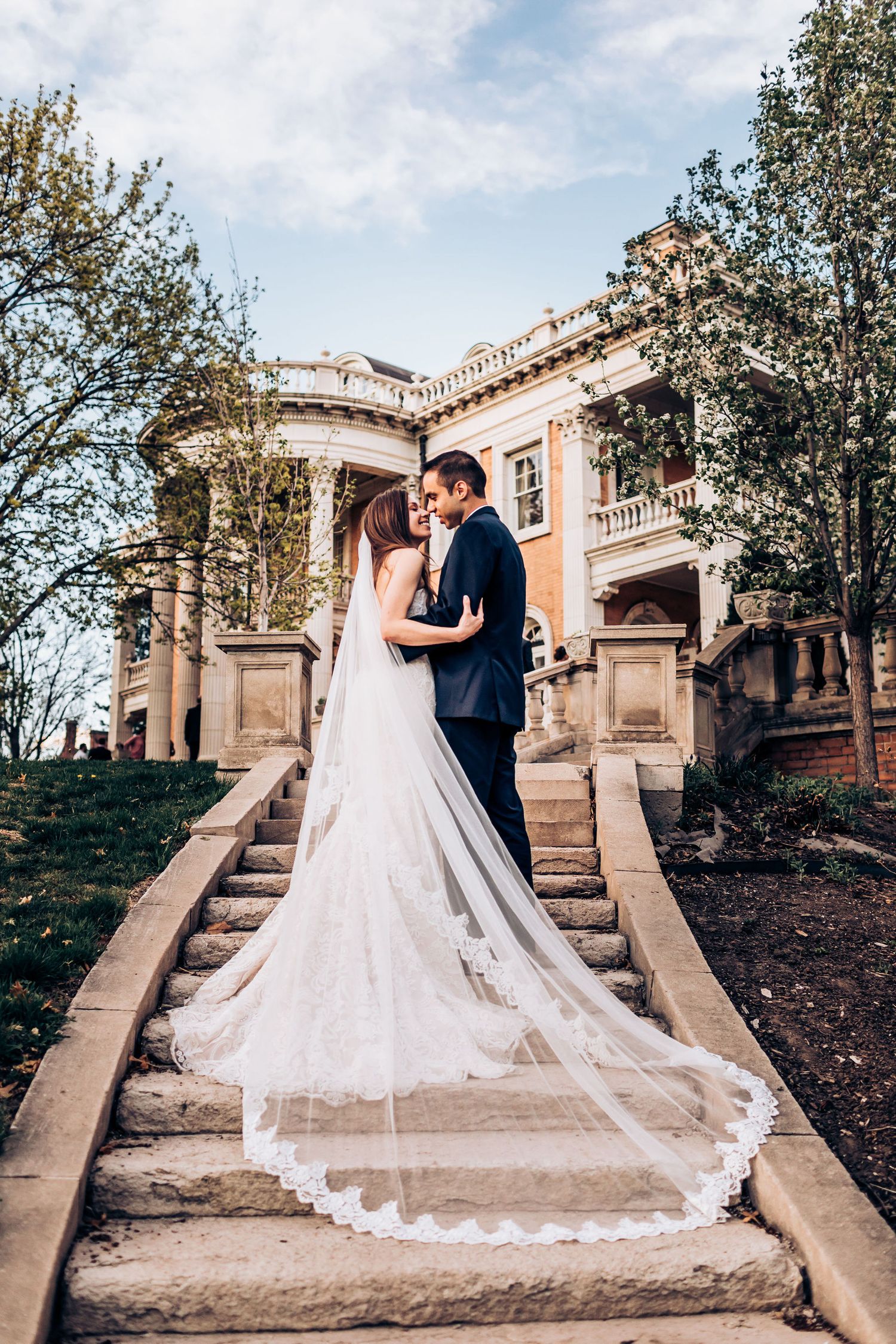Wedding couple poses on stone staircase with dramatic cathedral veil spread across historic mansion steps.