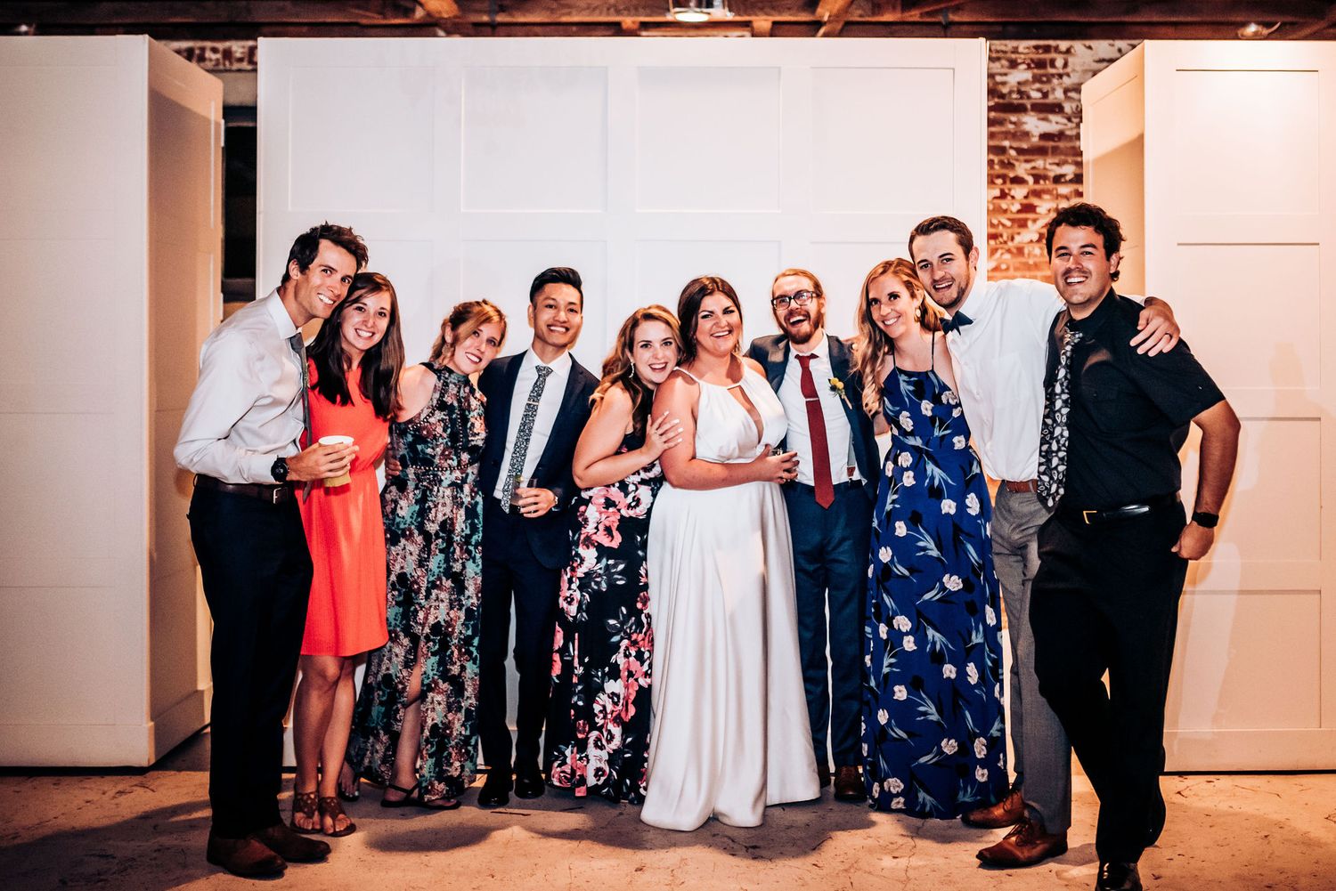 Wedding party poses together in formal attire against a white wall with exposed brick details.