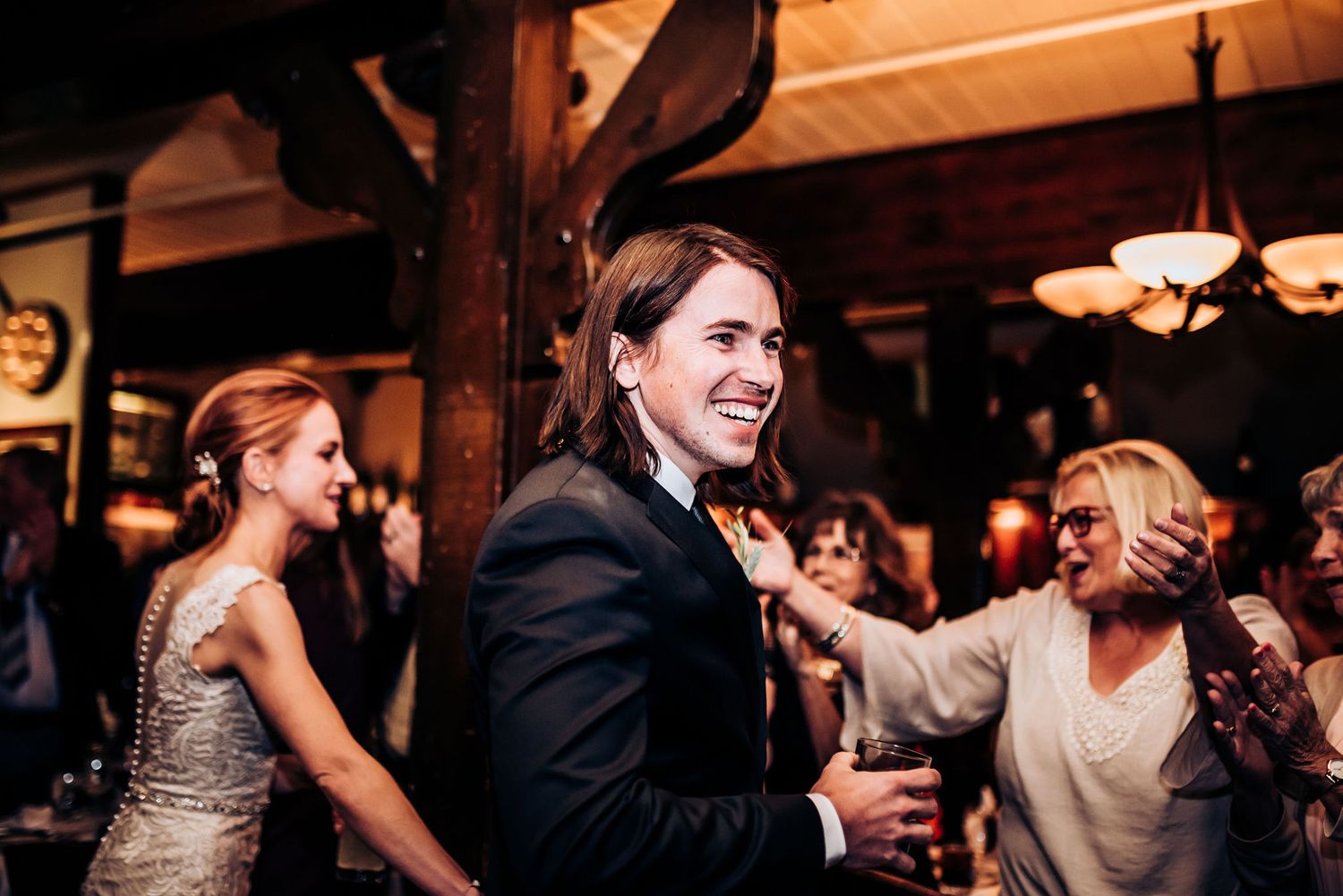 Wedding guests dance and celebrate in a dimly lit venue with wooden beams.