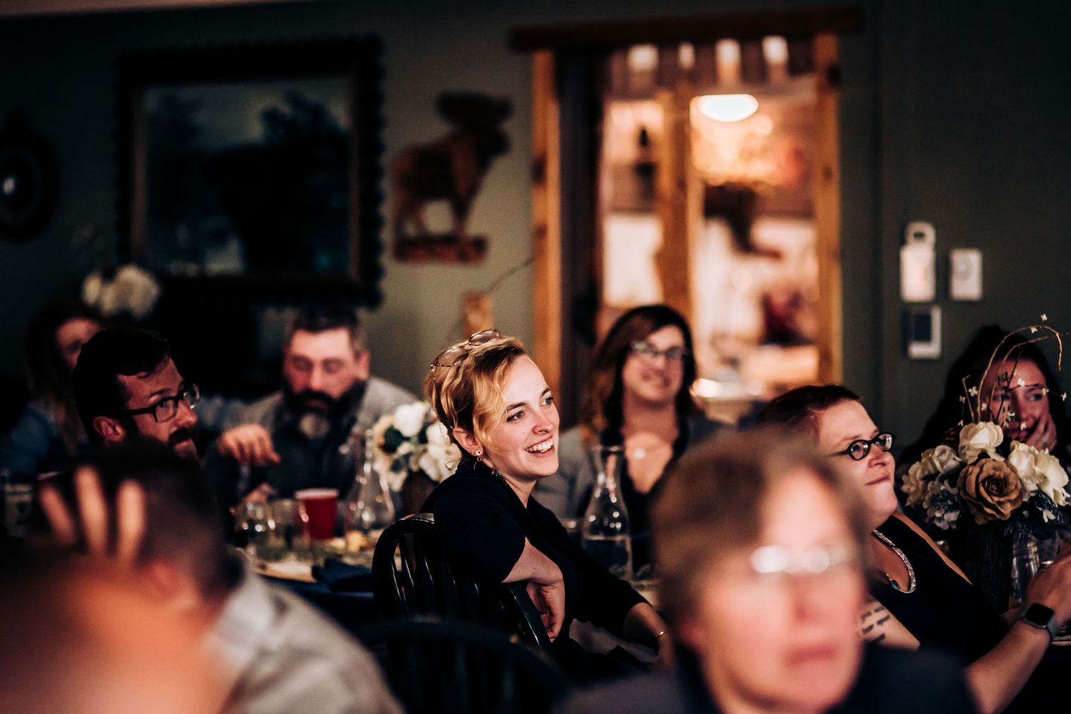 Wedding reception guests watch entertainment in a dimly lit rustic venue with floral centerpieces.