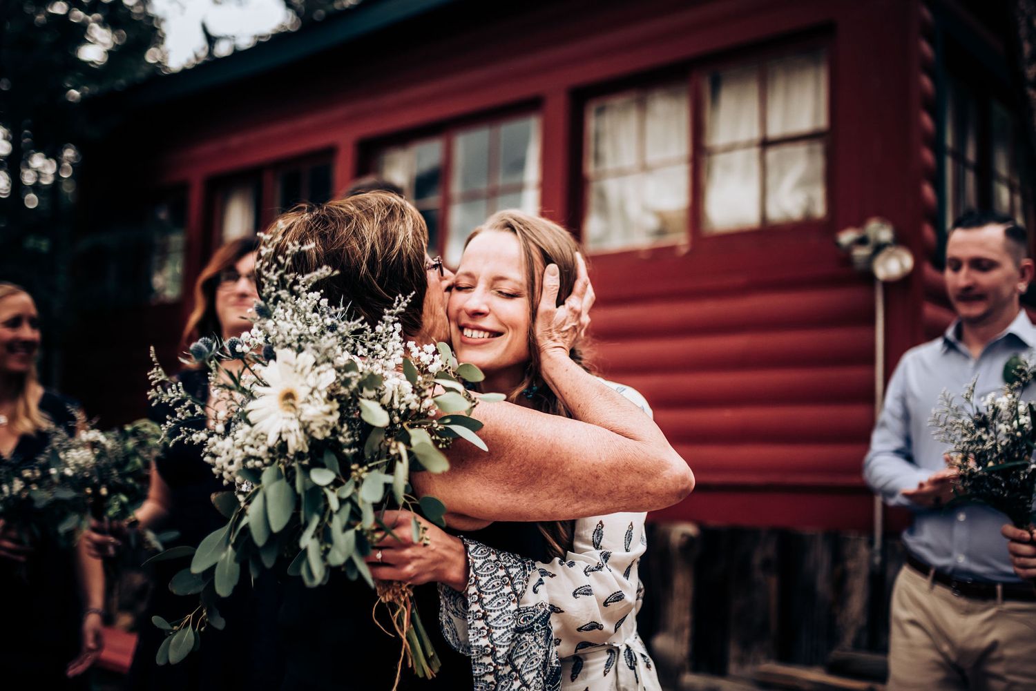 Friends share warm hugs while holding white flower bouquets during an outdoor celebration.