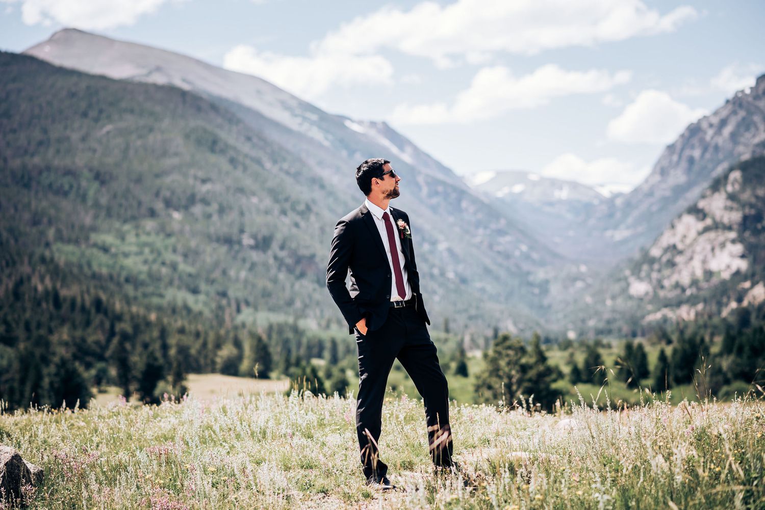 Groom in black suit stands alone in mountain meadow with dramatic valley vista behind him.