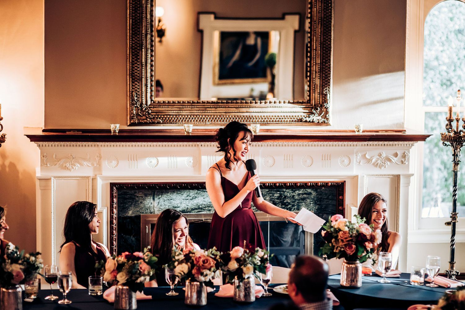 A speaker addresses wedding guests during a reception in an elegant room with a fireplace.
