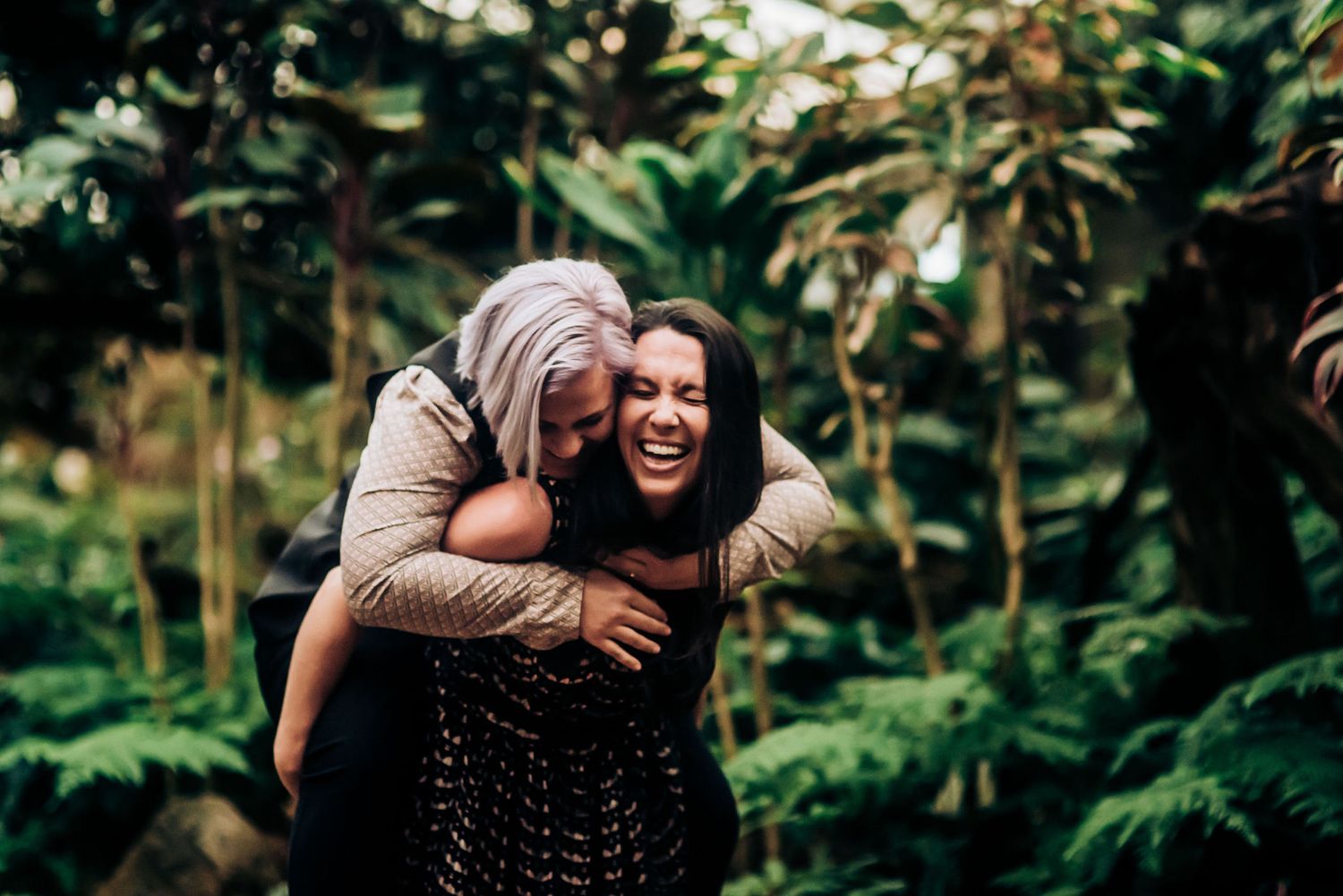 Two people share a joyful piggyback moment surrounded by lush greenery in a garden setting.
