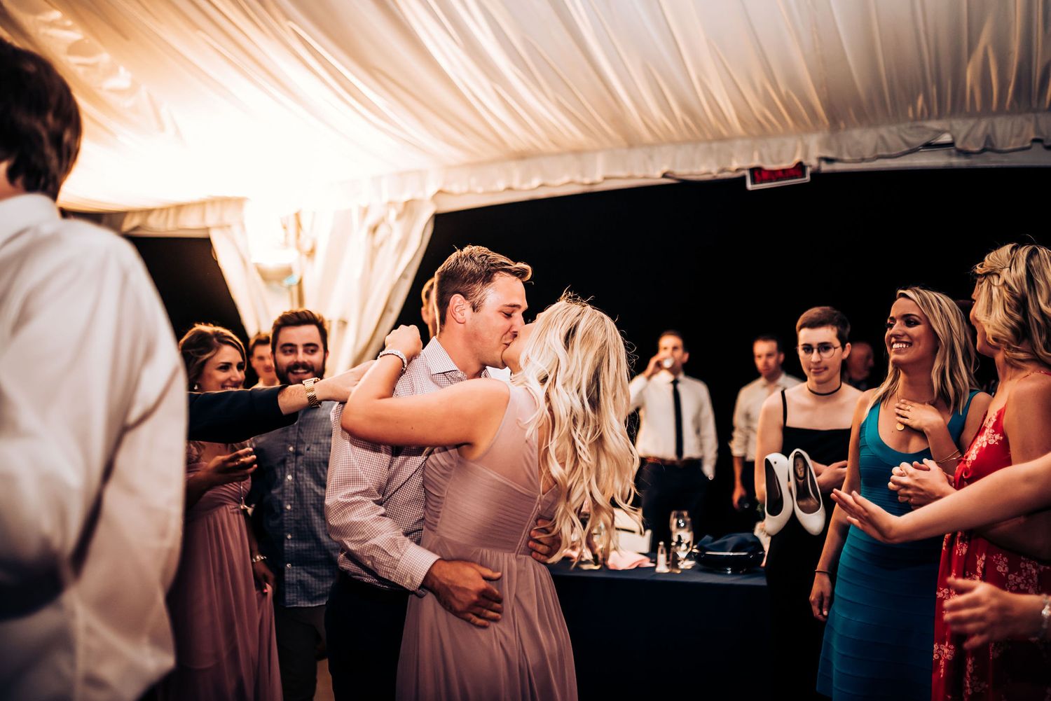 Couples slow dance surrounded by wedding guests under a white tent canopy.