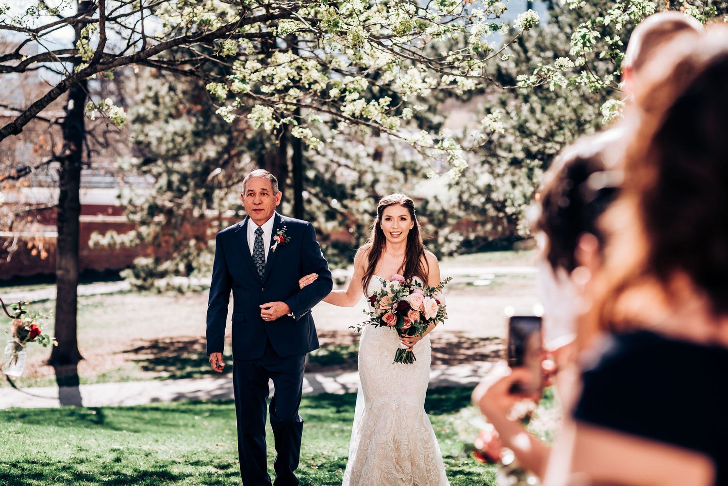 Spring wedding ceremony processional with blooming trees and guests taking photos.