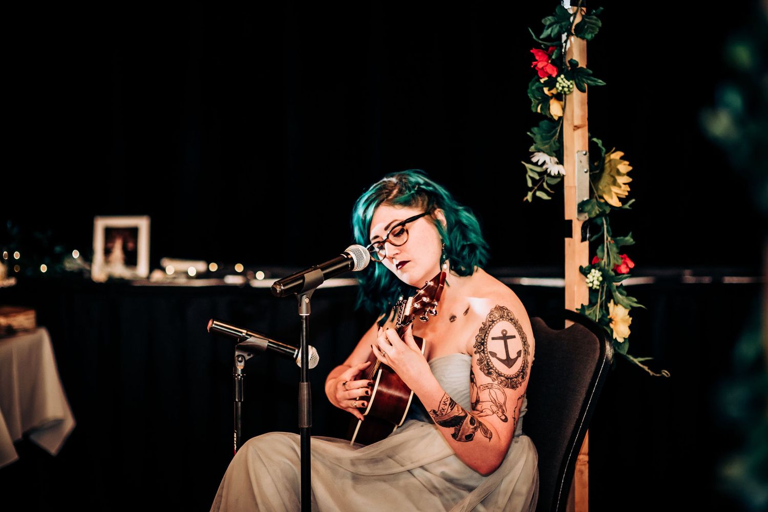 Musician with teal hair plays acoustic guitar while performing at wedding reception.