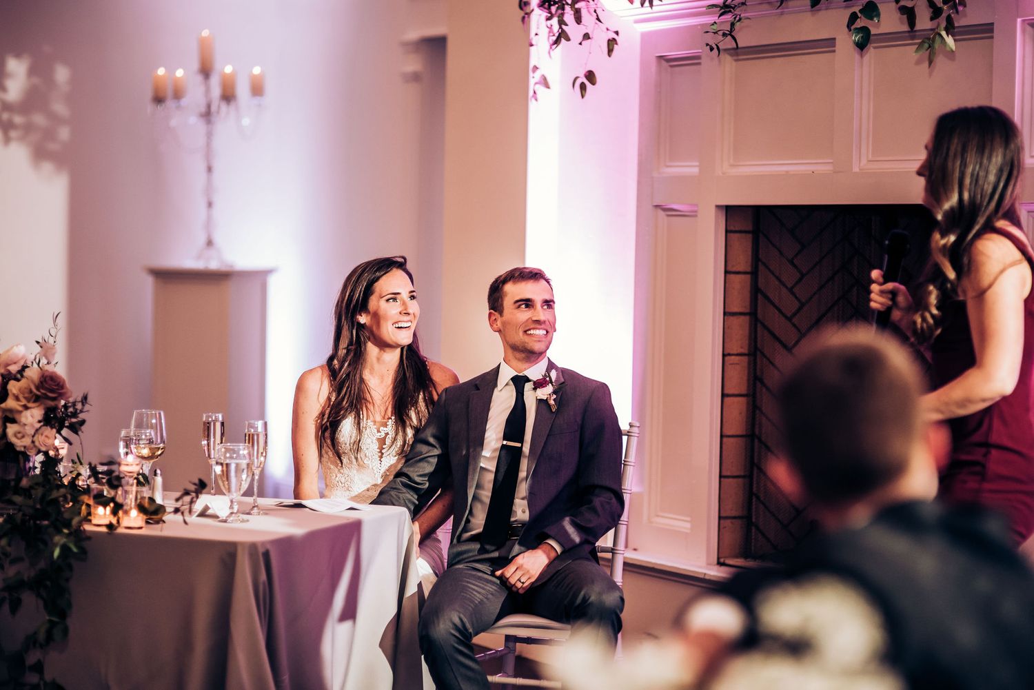 Wedding party members seated at head table while guest gives speech during evening reception.