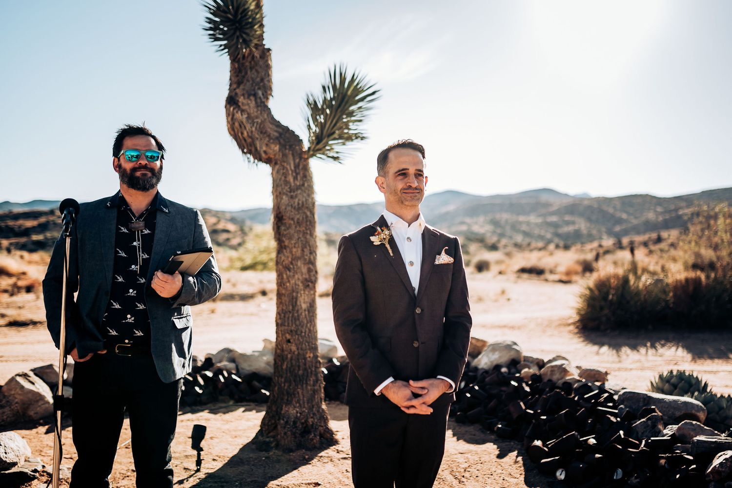 Two people stand in a desert landscape with Joshua trees during a sunny day.