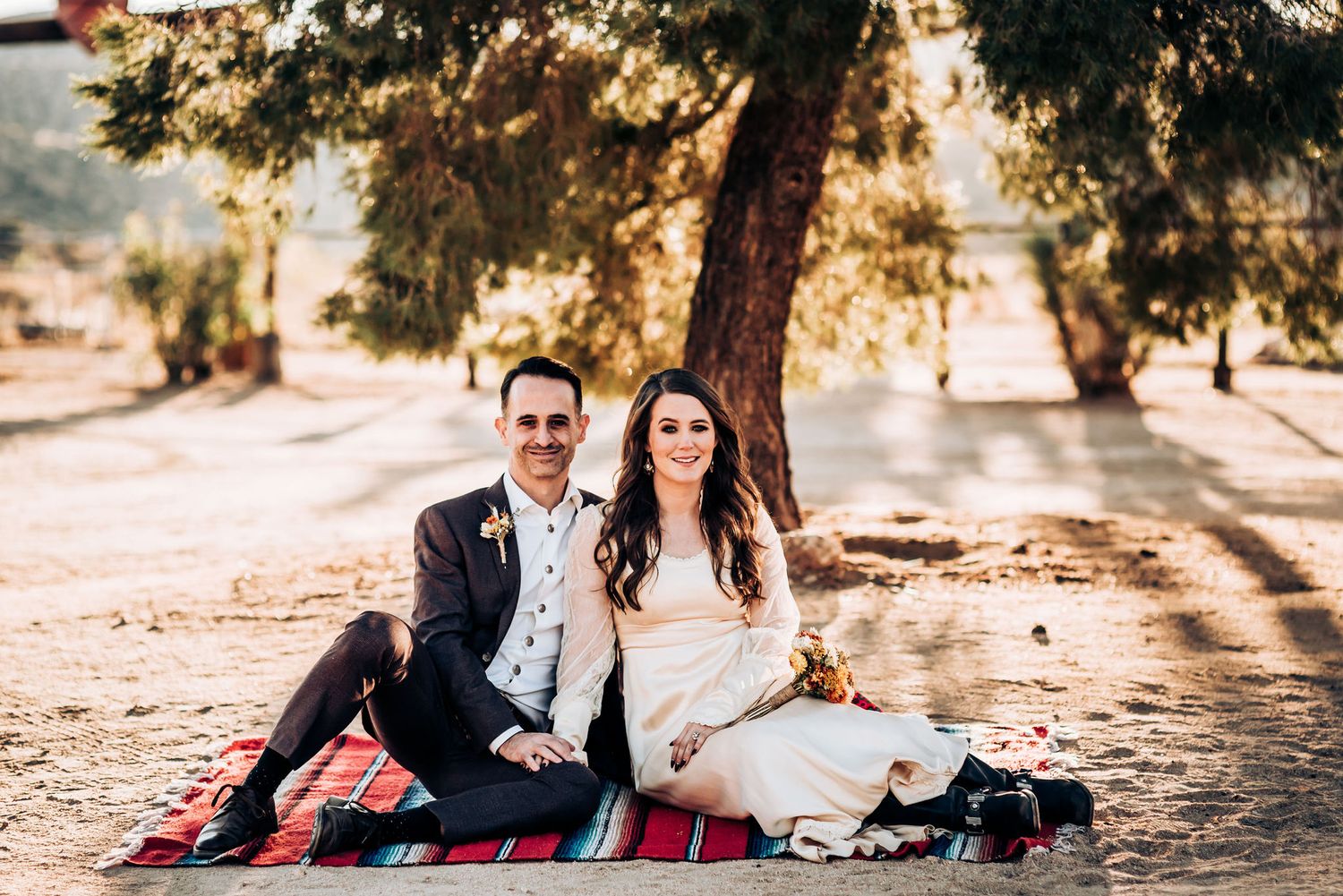 A couple sits on a red blanket under a tree during a romantic outdoor photoshoot at golden hour.