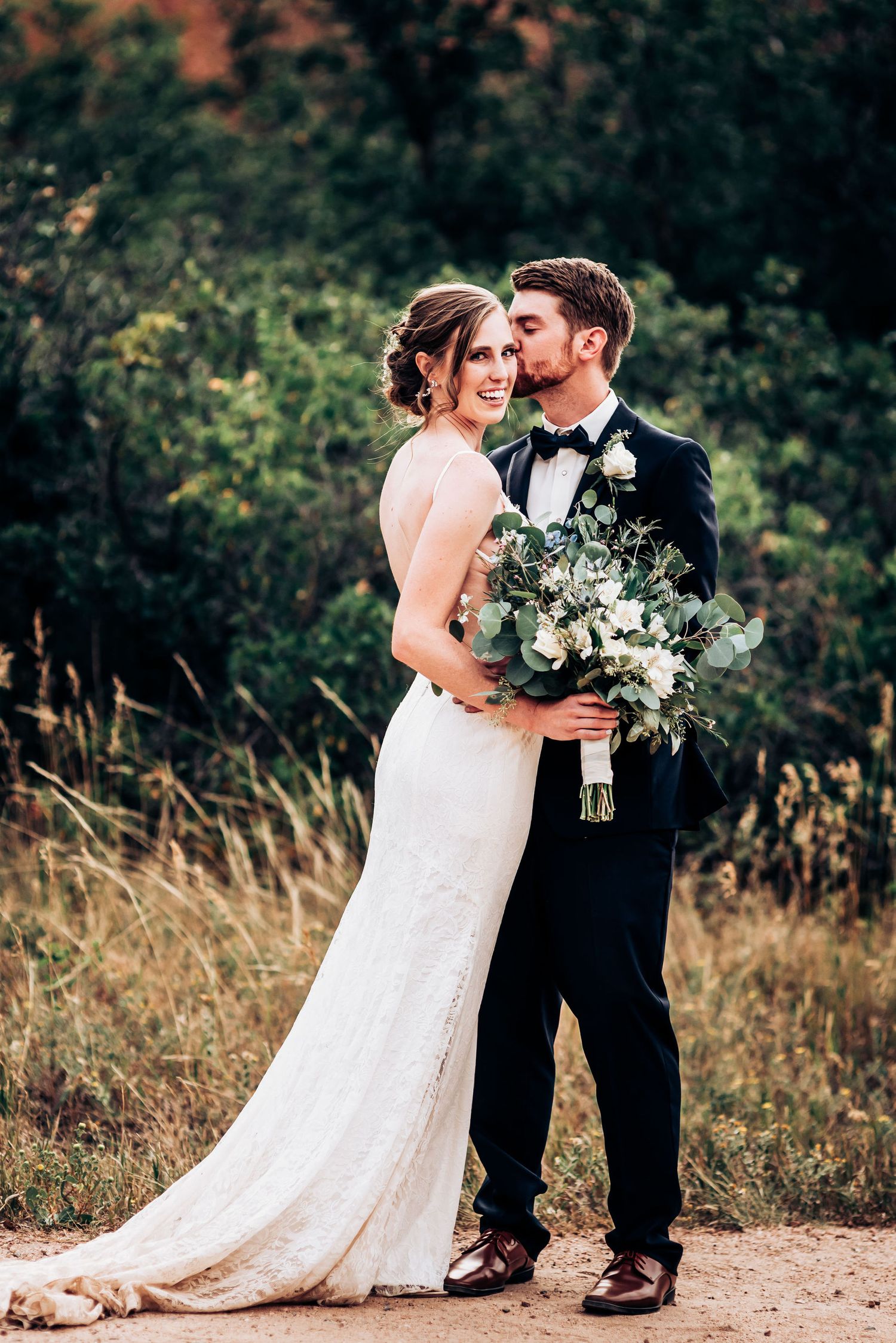 A couple embraces on a dirt path while holding an elegant white and green bouquet.