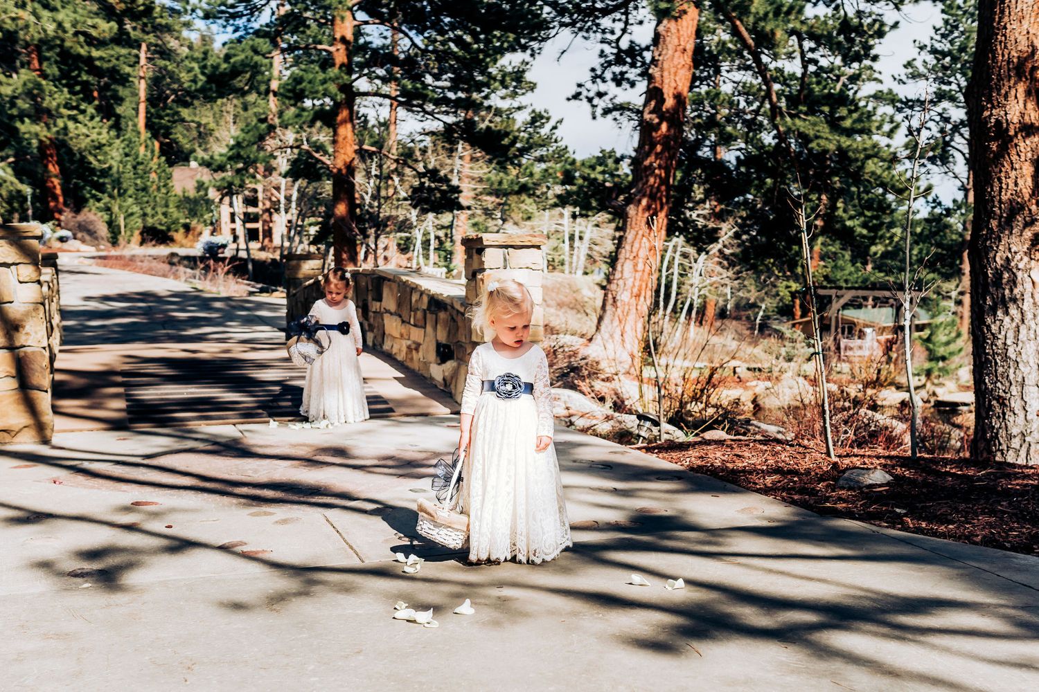 Flower girl in white dress walks down sunny outdoor pathway during wedding ceremony.