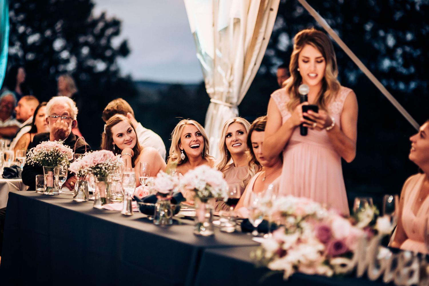 Wedding party members at a reception dinner table react to speeches under evening lighting.