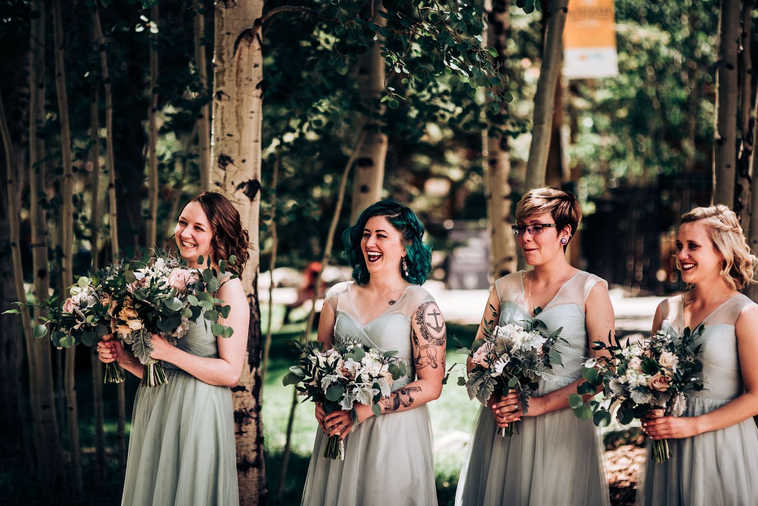 Four bridesmaids in sage green dresses hold white floral bouquets while standing in a wooded outdoor setting.