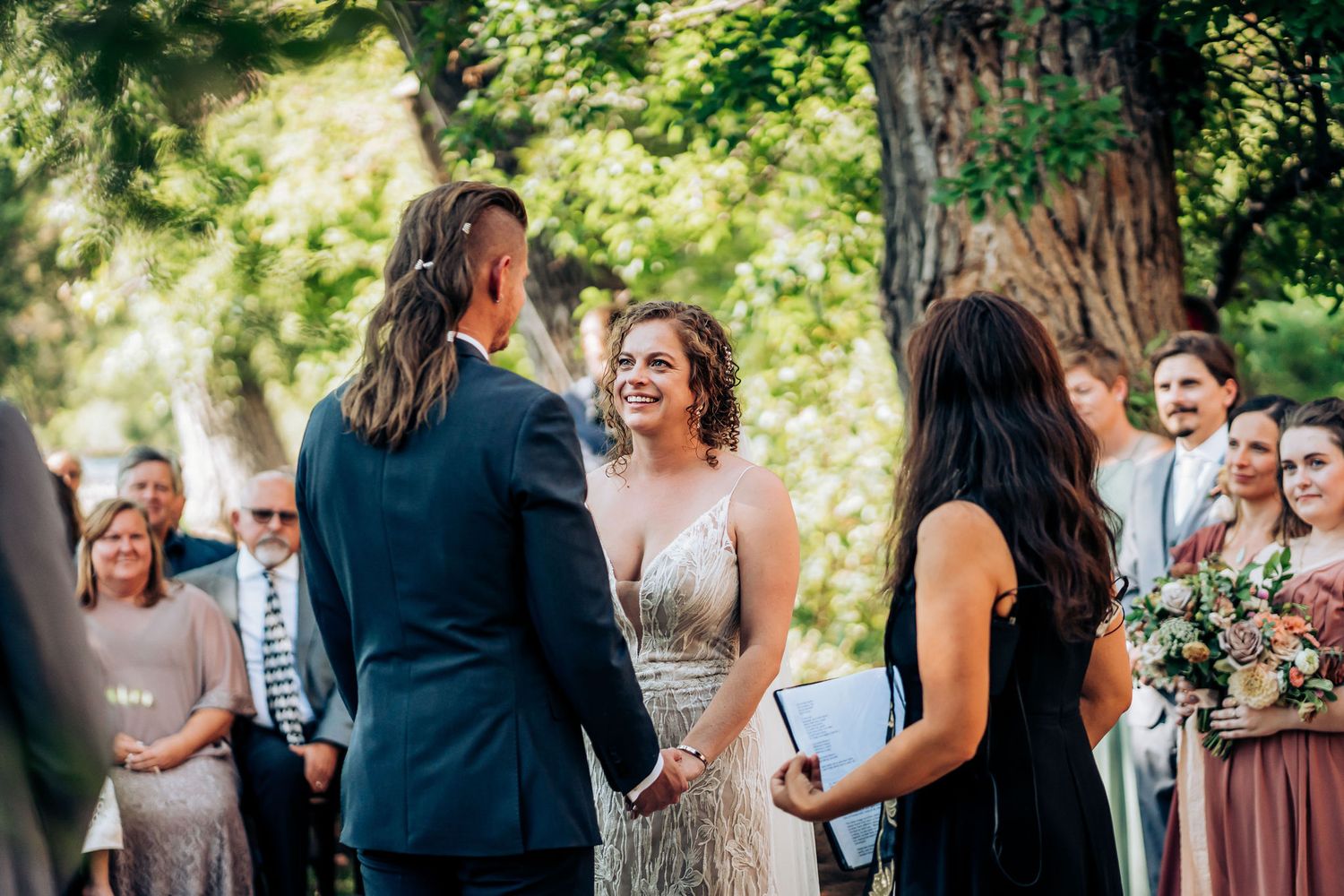 Outdoor wedding ceremony with smiling attendees under natural tree canopy.
