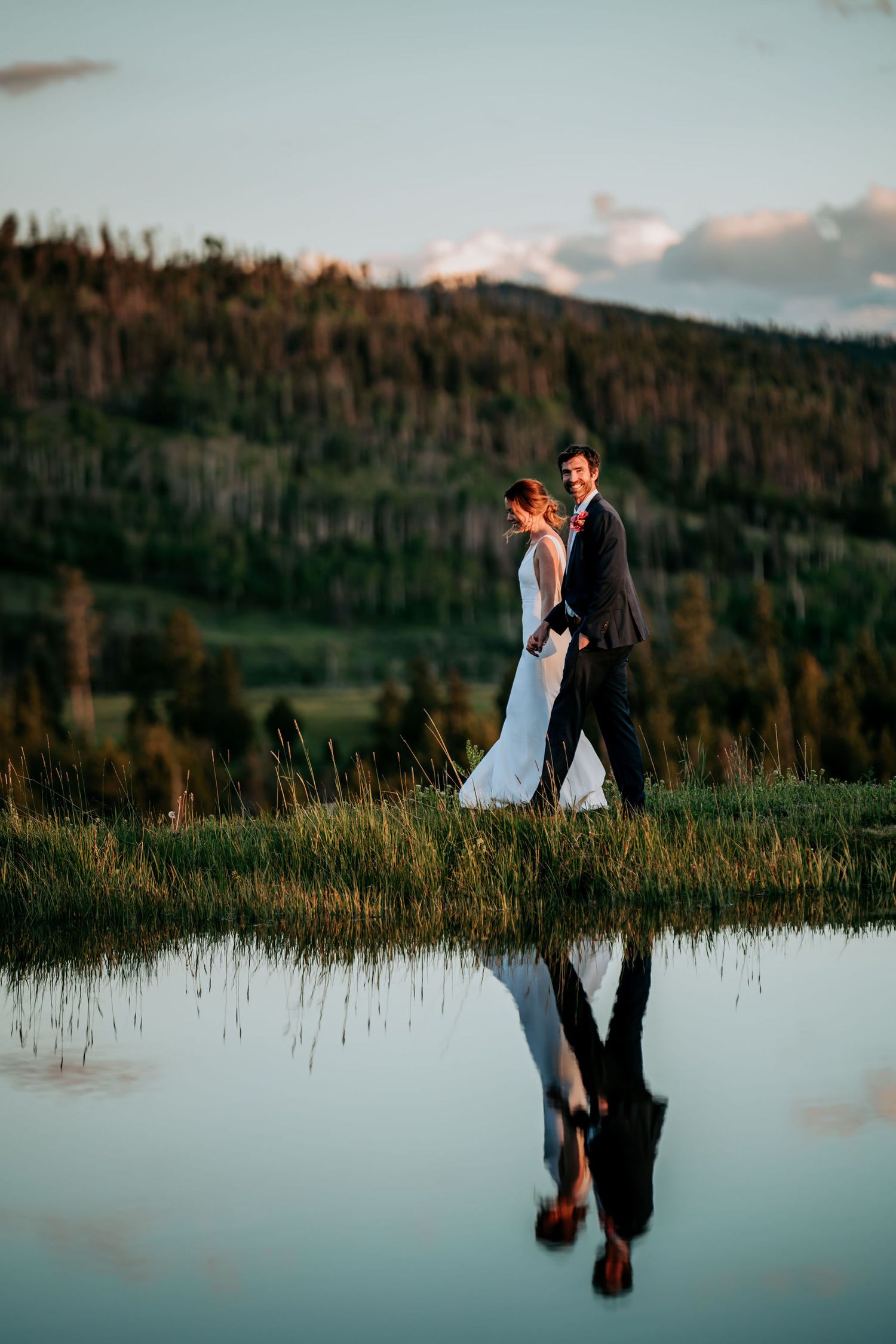 Wedding couple stands at mountain lake's edge with perfect reflection at sunset.