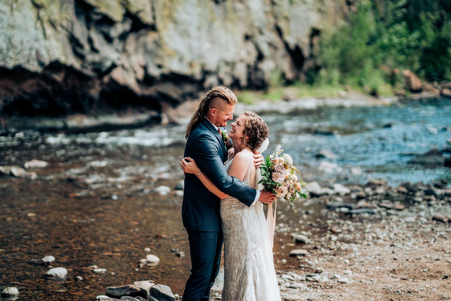 Romantic wedding portrait with couple embracing near flowing river and rocky cliffs.