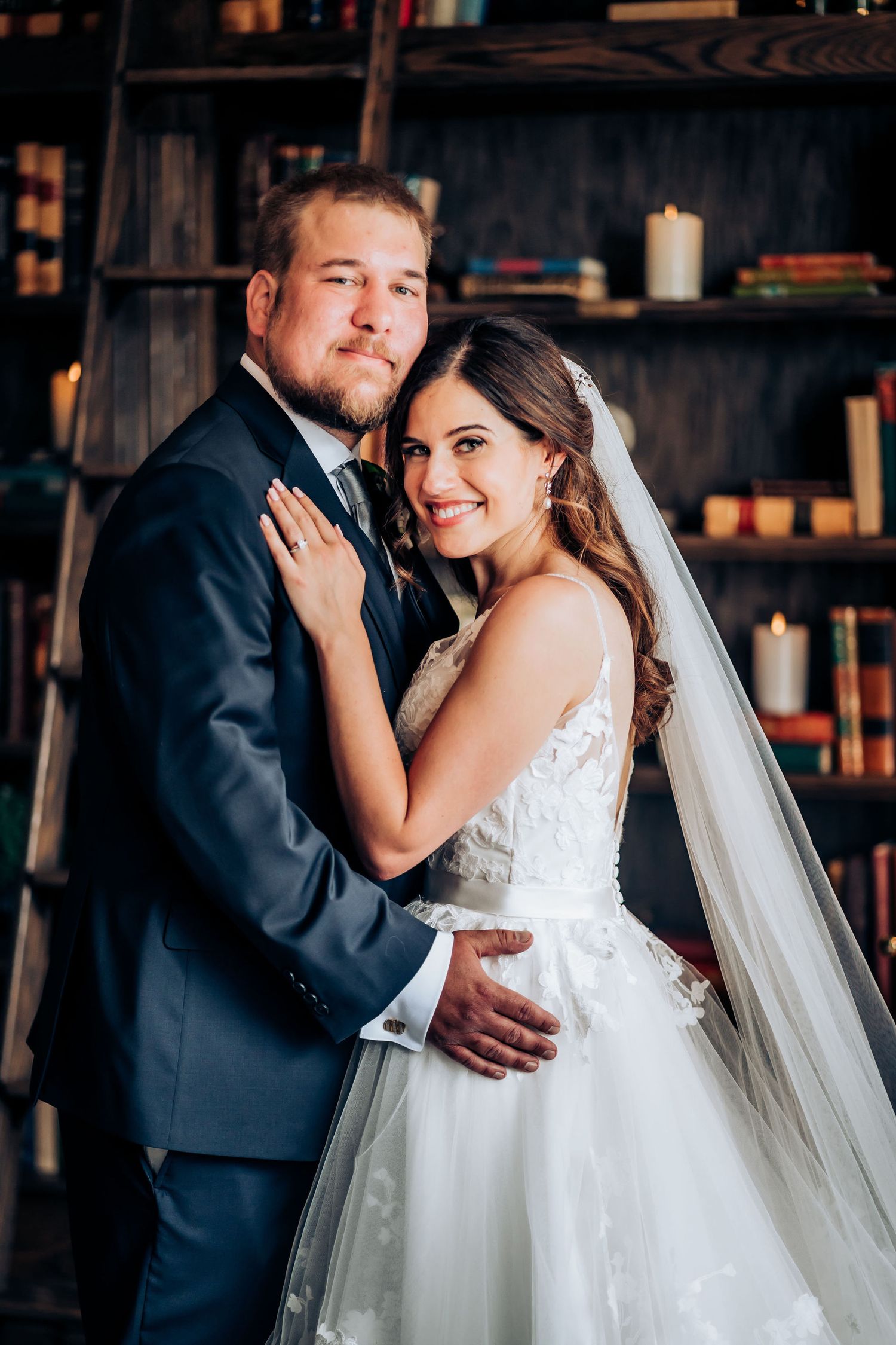 Newlyweds share intimate moment in rustic library setting with wooden shelves and candles.
