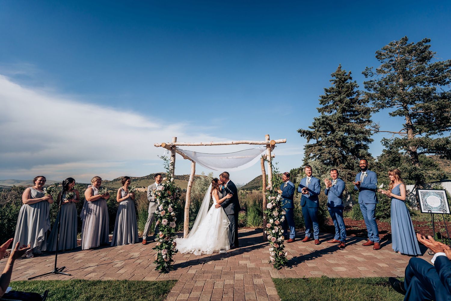 Panoramic view of wedding ceremony under rustic wooden arch with wedding party and guests.