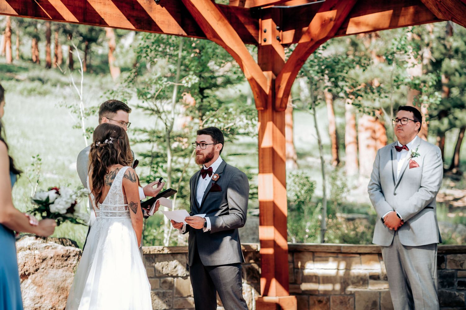 An outdoor wedding ceremony takes place under a wooden pavilion surrounded by forest.