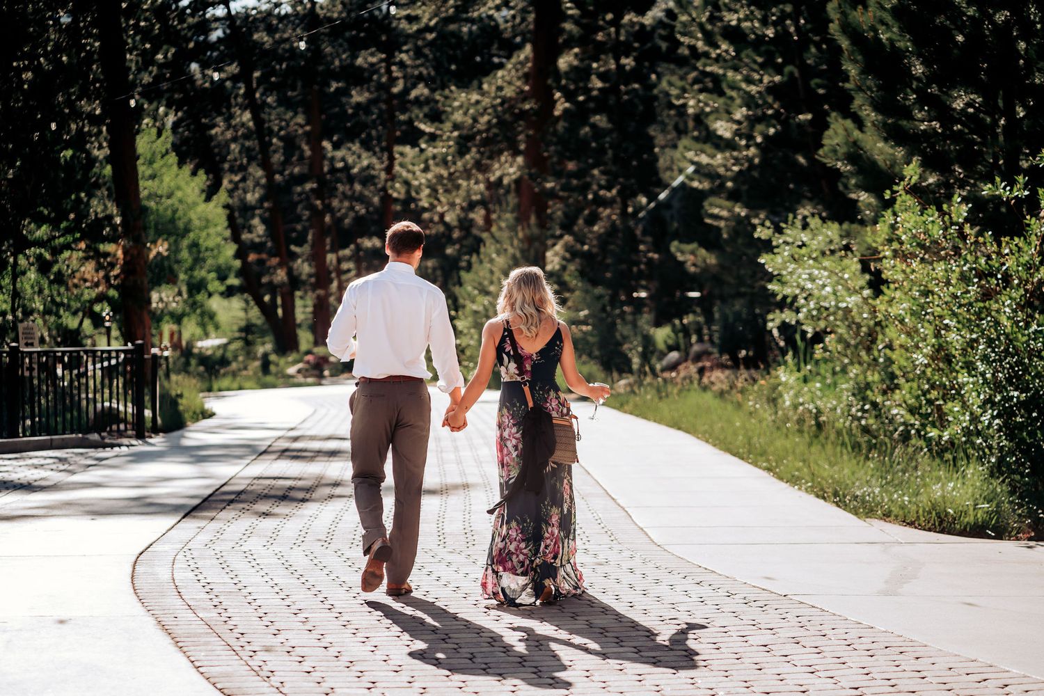 A couple walks hand in hand down a scenic paved path surrounded by trees.