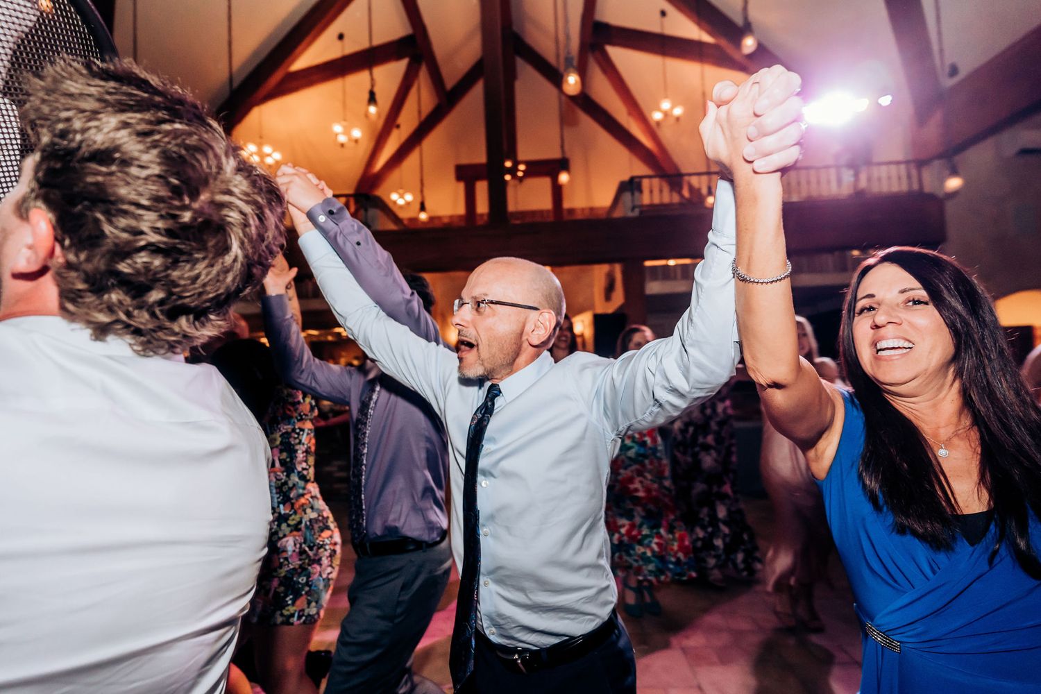 Wedding guests raise their arms in celebration during a lively dance party in a rustic venue.