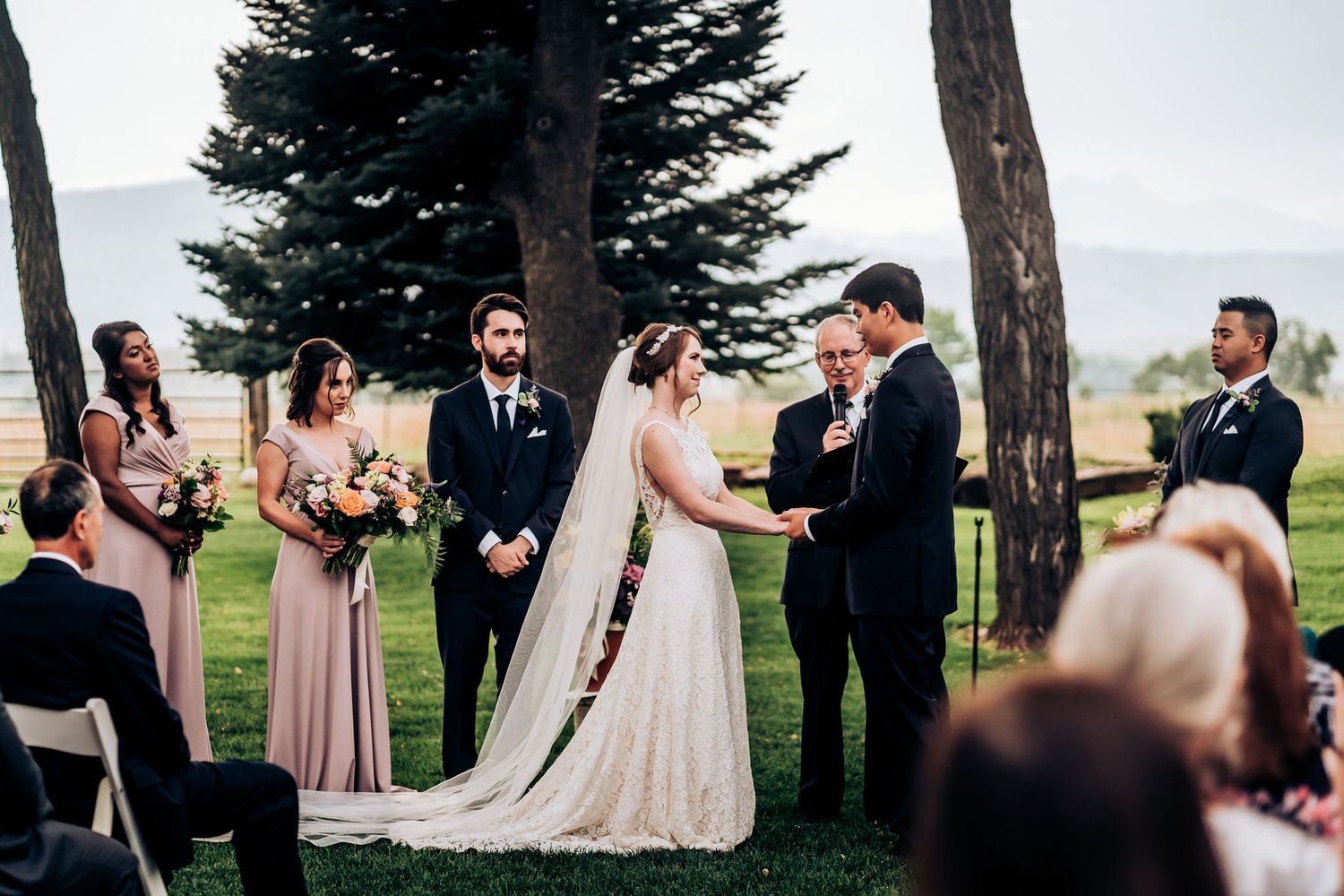 Intimate outdoor wedding ceremony among evergreen trees with guests seated on white chairs.