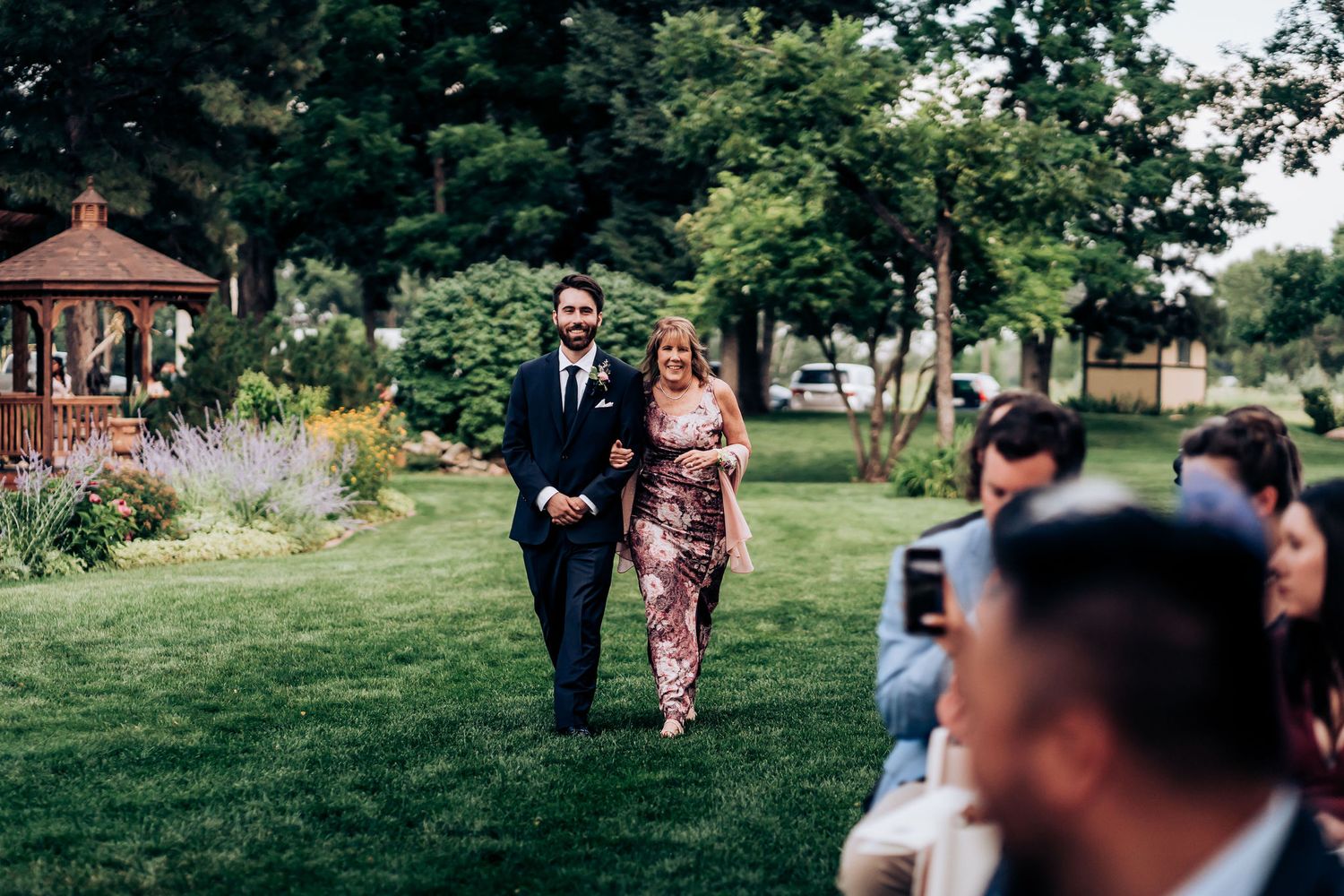 Wedding processional on a landscaped lawn with guests seated outdoors.