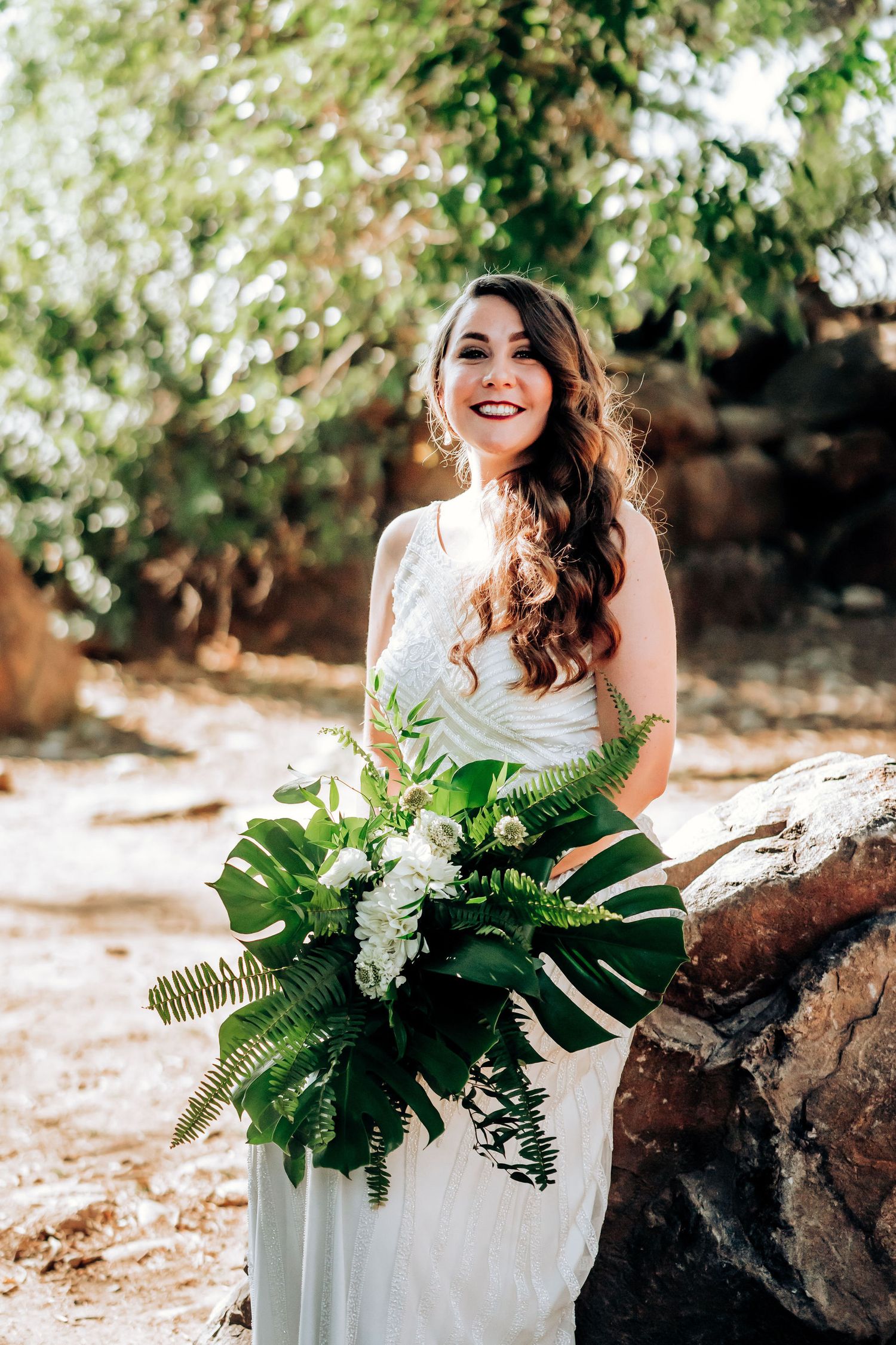 An elegant wedding portrait featuring a white dress and tropical leaf bouquet in an outdoor setting.