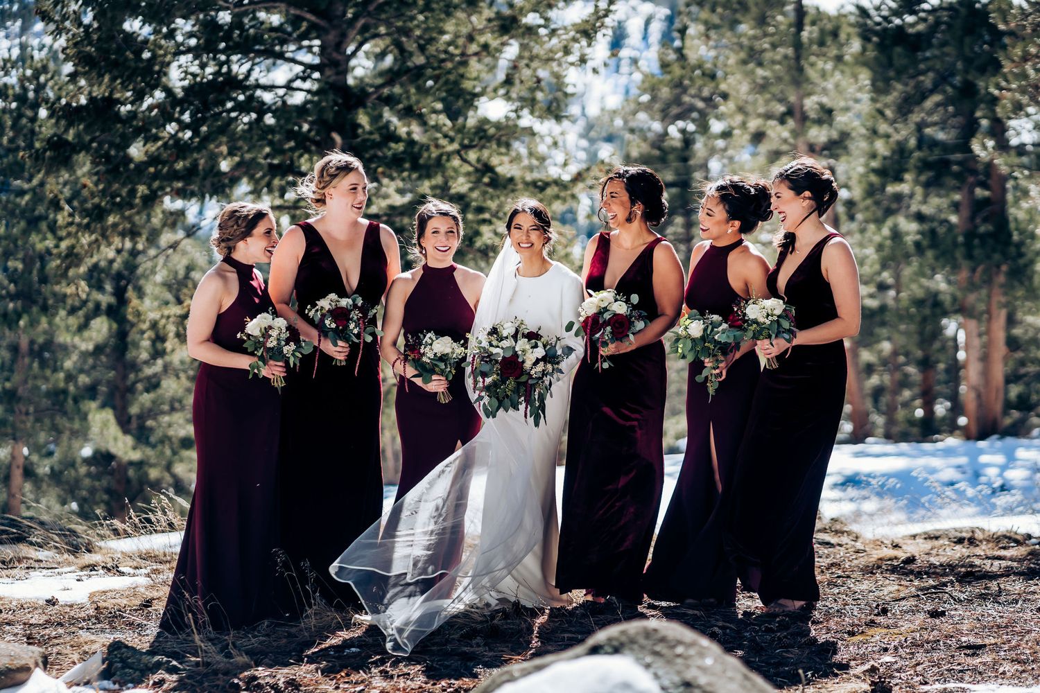 Wedding party in burgundy dresses poses on snowy mountain terrain with evergreen forest background.