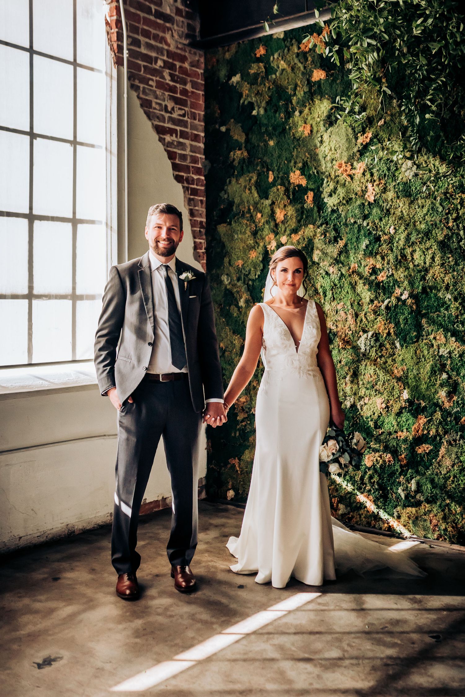 Elegant couple in wedding attire holding hands in front of a brick wall with climbing vines.