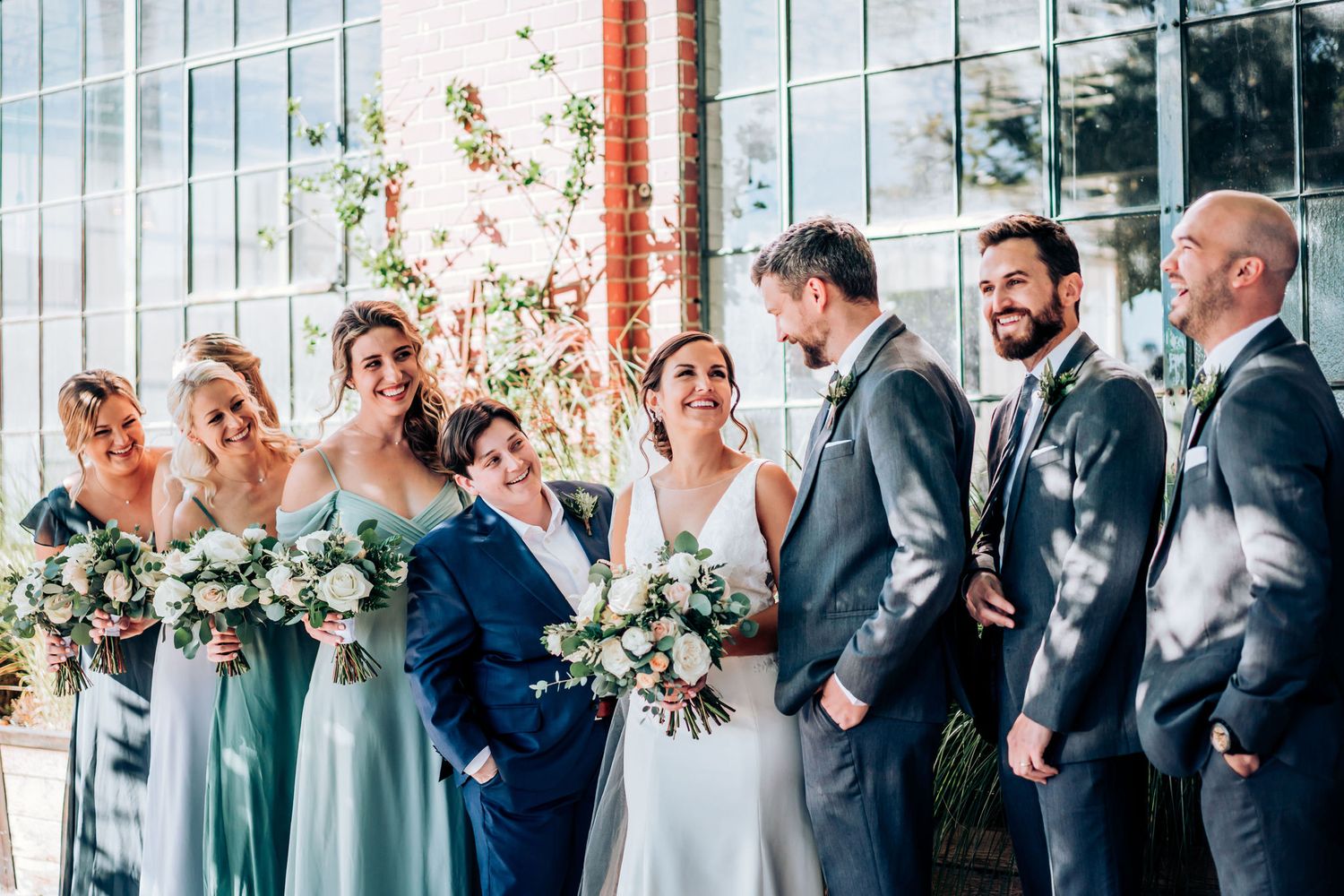 Wedding party with bridesmaids in sage dresses gather near large windows in modern venue.