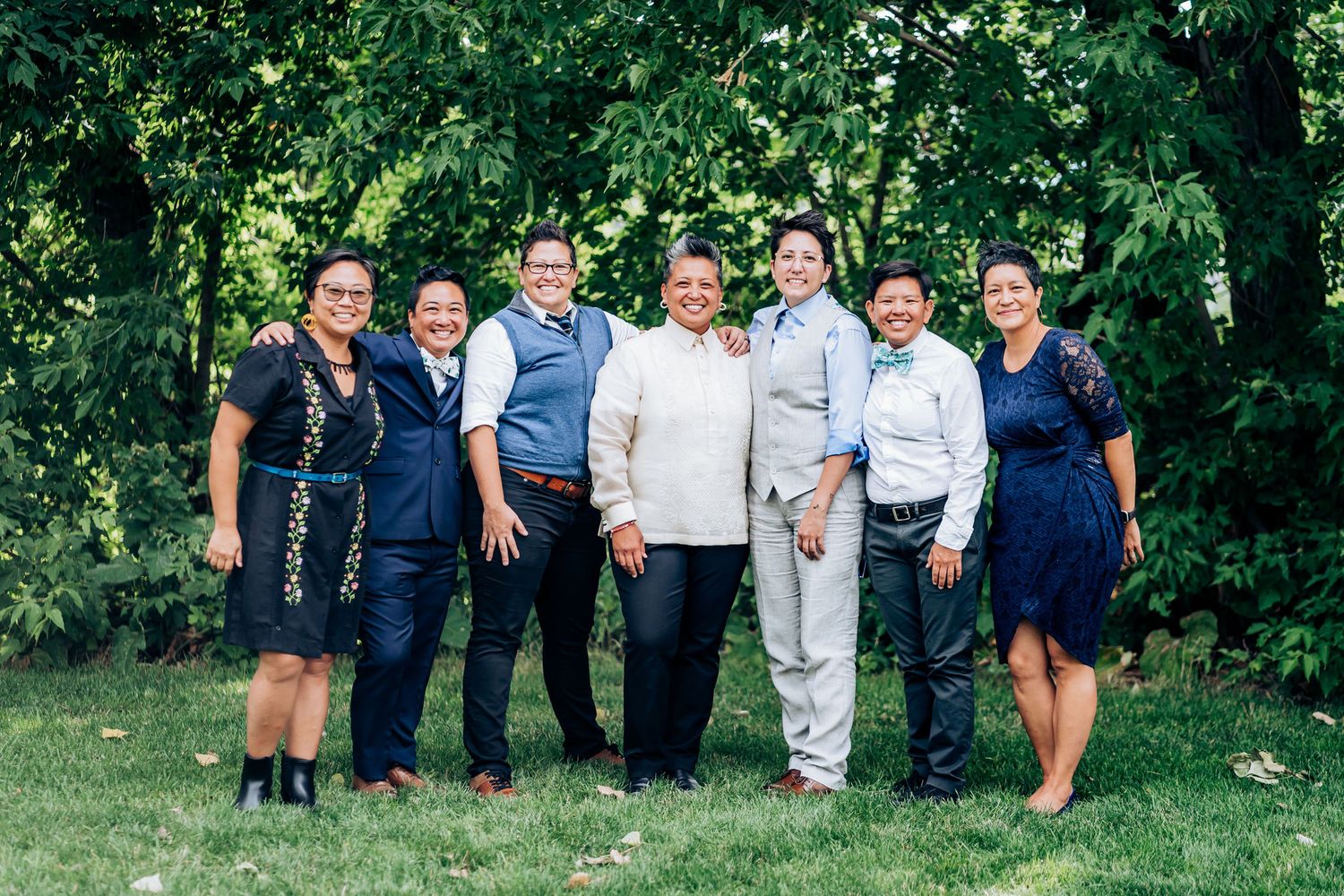 Group photo of wedding party members in formal attire standing together on grass with trees behind them.