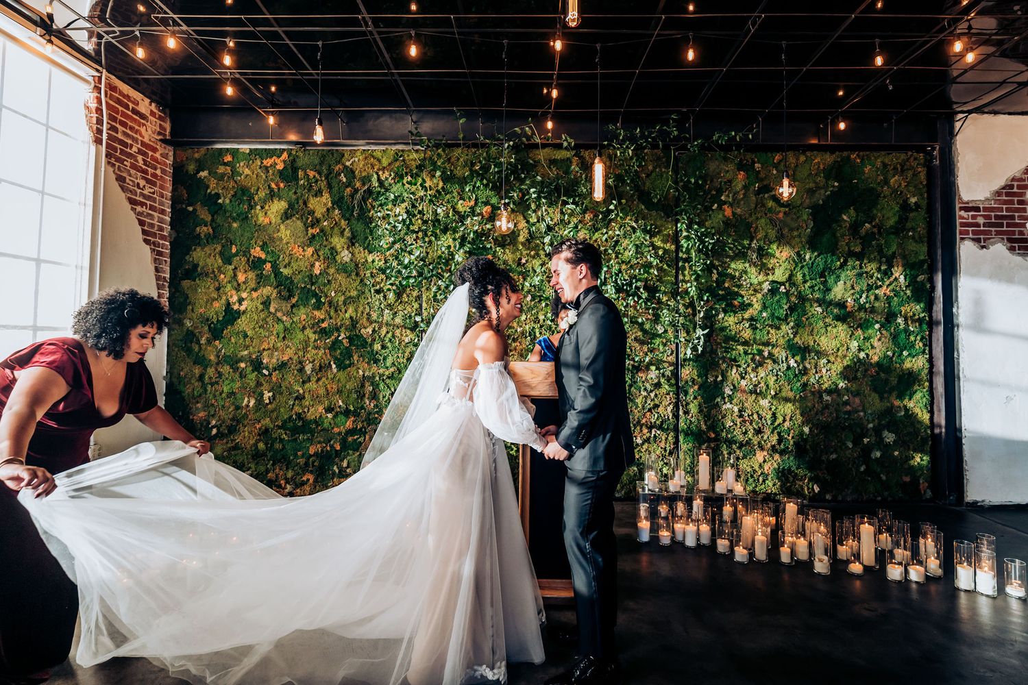 Wedding ceremony with attendants adjusting bride's flowing veil against greenery backdrop.