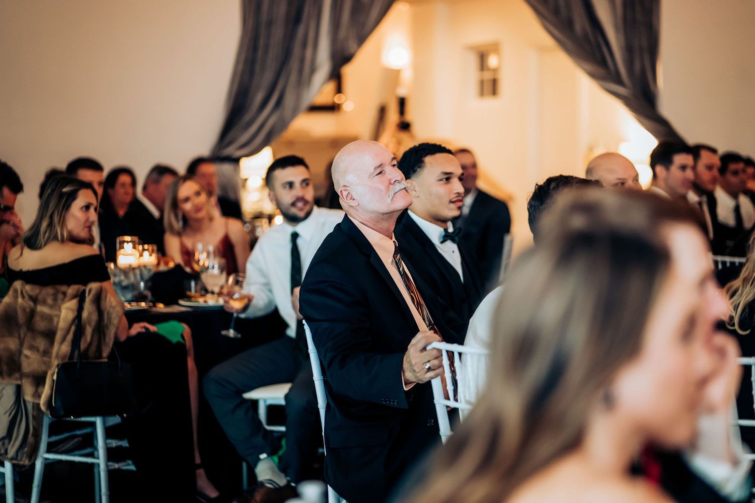 Wedding guests seated in formal attire during an indoor reception with dramatic lighting.