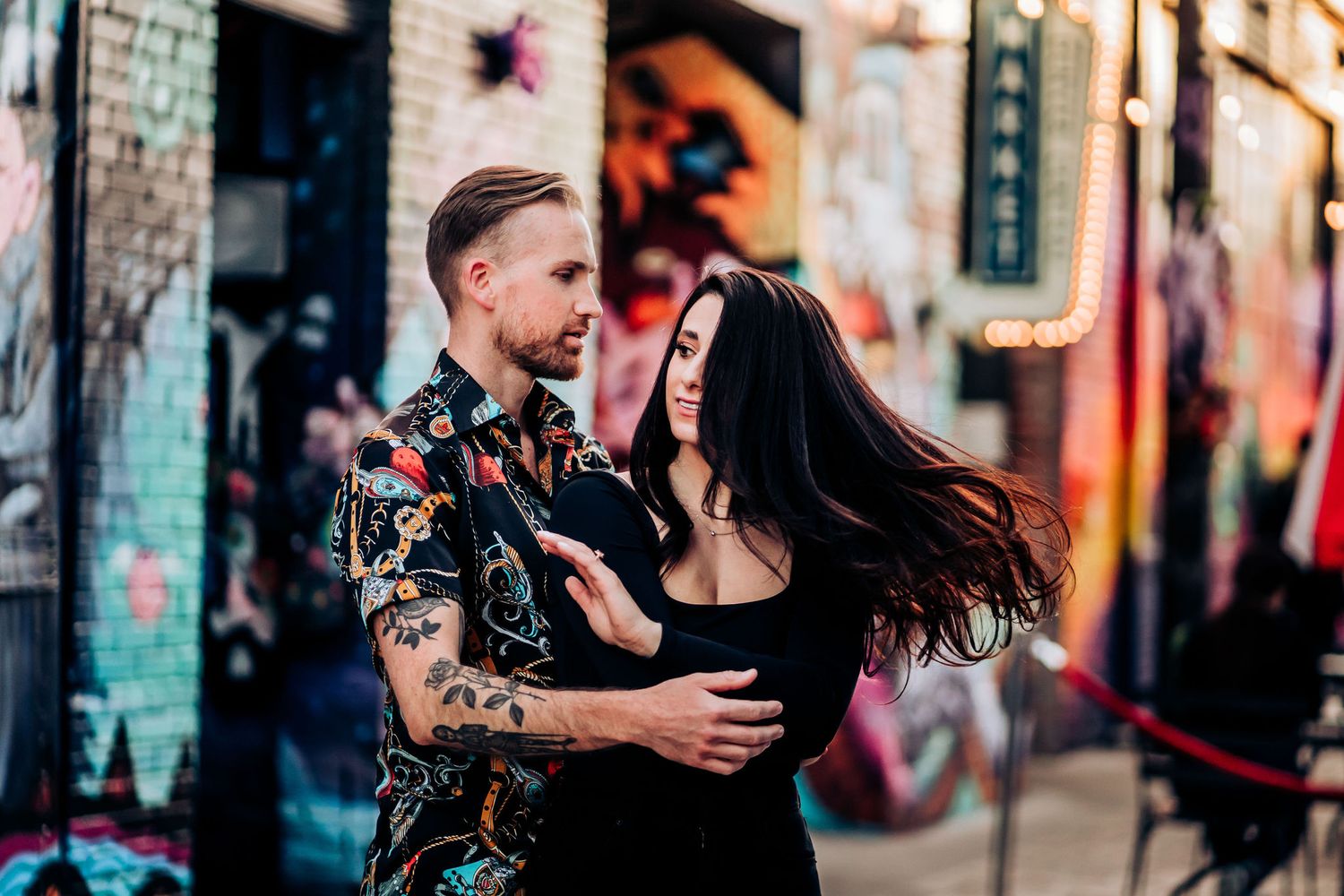 A couple embraces in front of vibrant street art while one's hair flows in the wind.