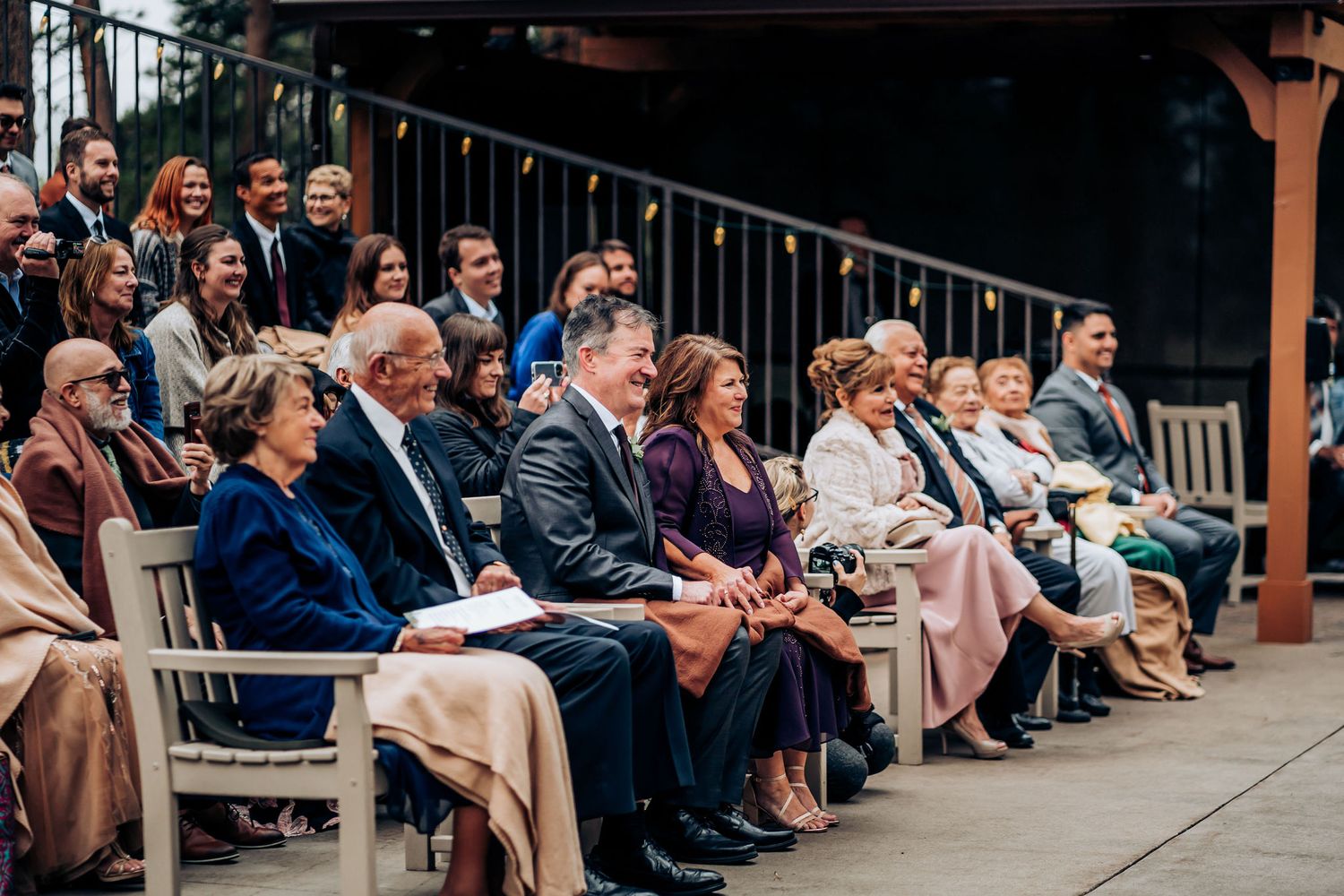 Wedding guests seated in white chairs watching ceremony under covered outdoor venue.