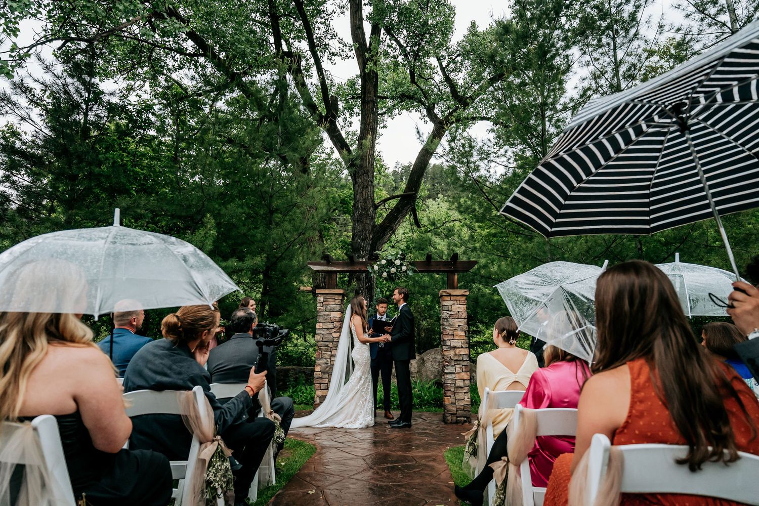 A rainy outdoor wedding ceremony with guests holding clear umbrellas amidst lush greenery.