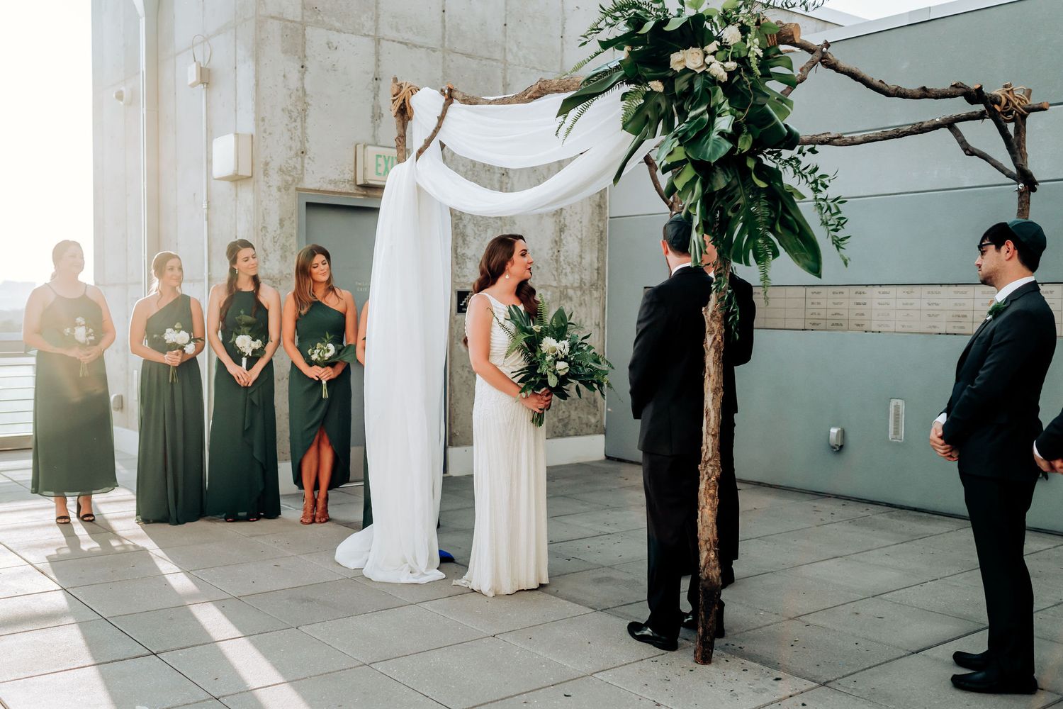 Modern outdoor wedding ceremony with bridesmaids in emerald green dresses.