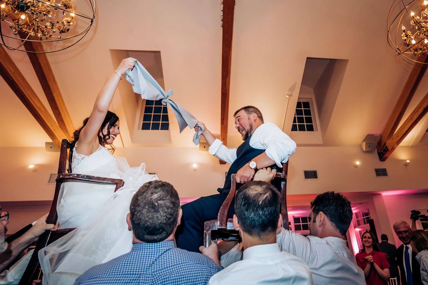 Guests lift bride and groom on chairs during lively wedding reception celebration.