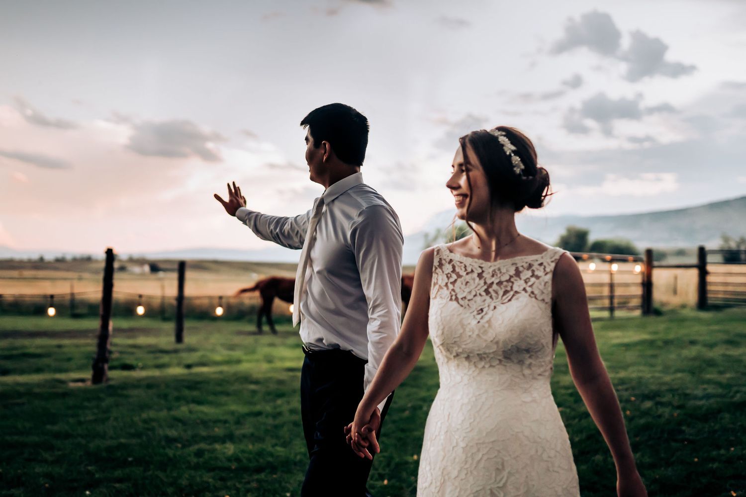 A bride in lace gown and groom in white shirt dance outdoors against a mountain landscape at sunset.