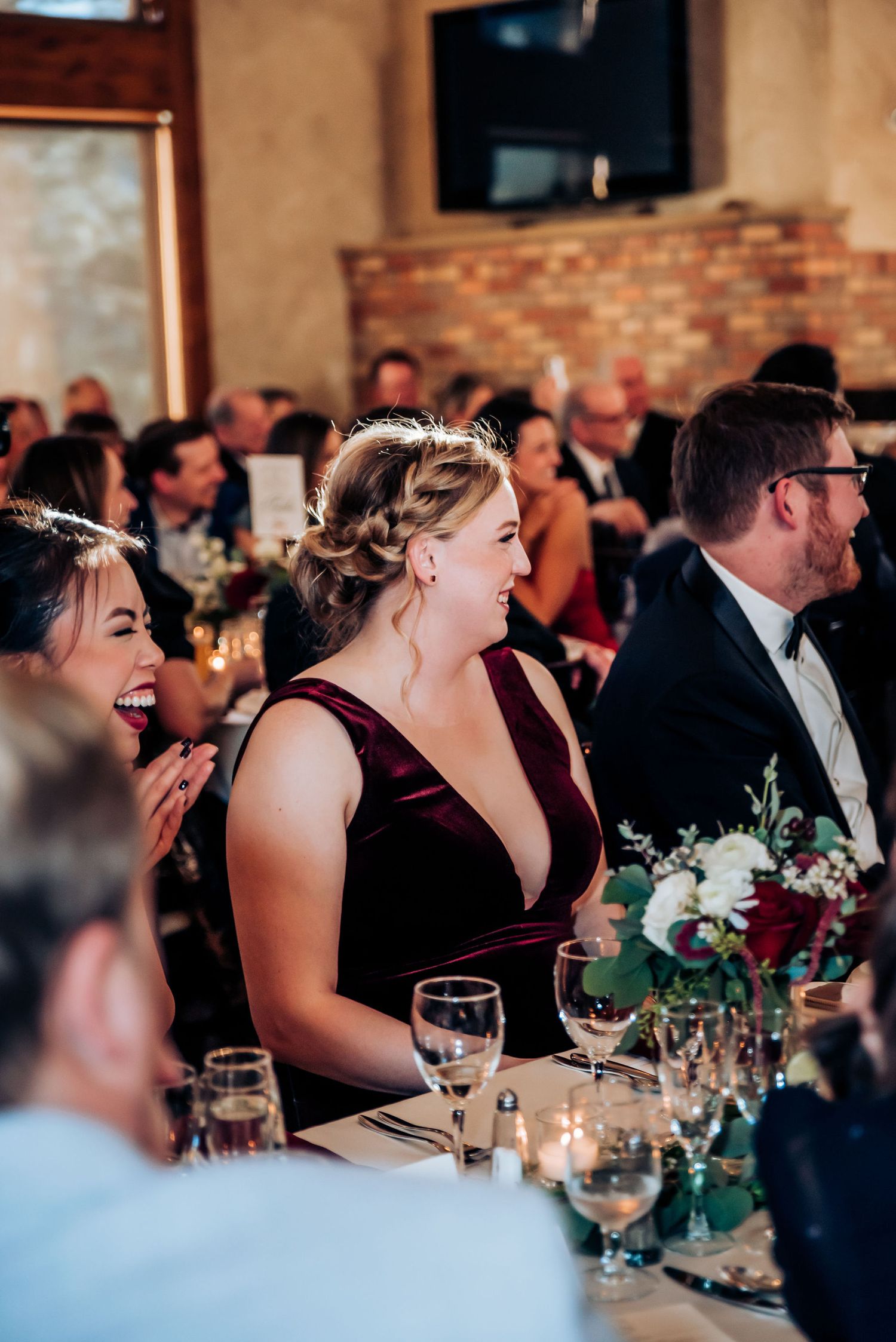 Wedding guests laugh and celebrate at a reception dinner with cozy brick walls and festive table settings.