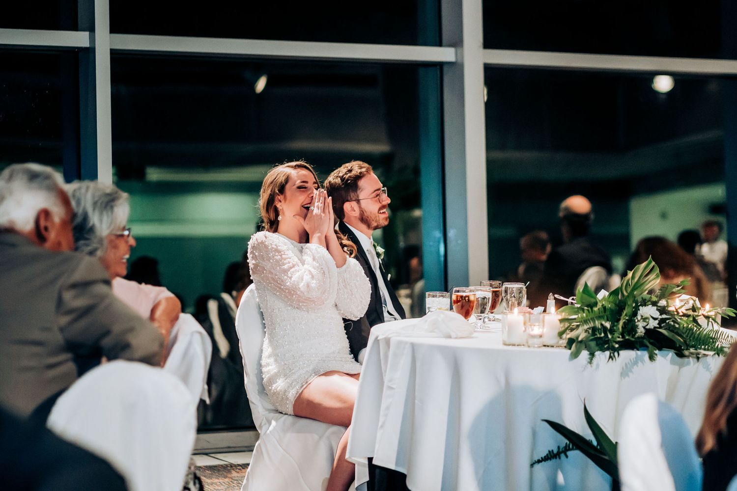 A couple celebrates joyfully at their reception table decorated with white linens and greenery.