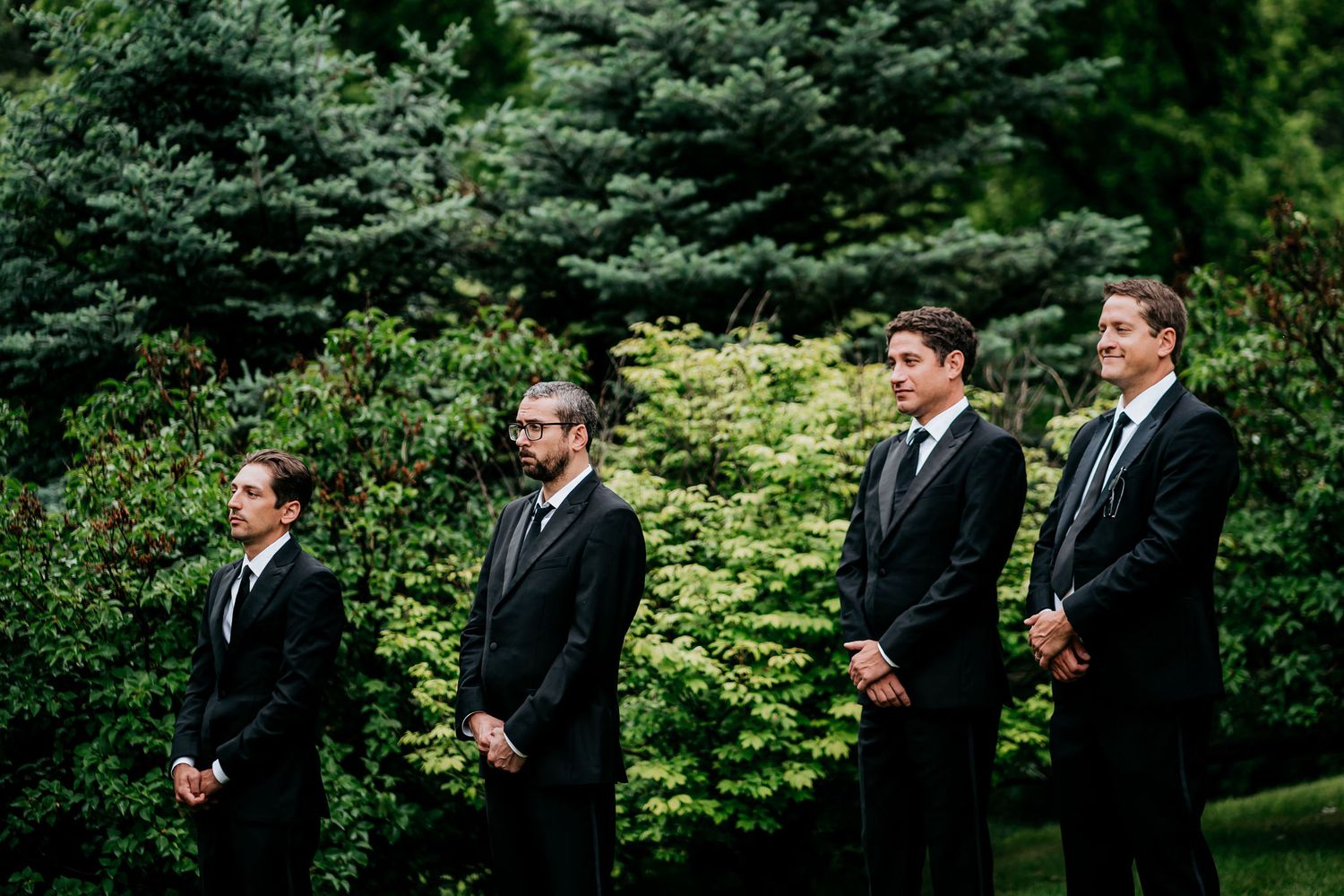 Groomsmen in black suits stand in a row during outdoor wedding ceremony.