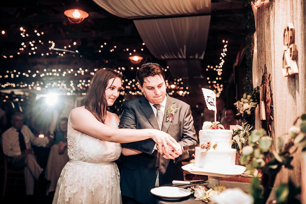 A couple cuts their wedding cake together at a reception under twinkling lights.