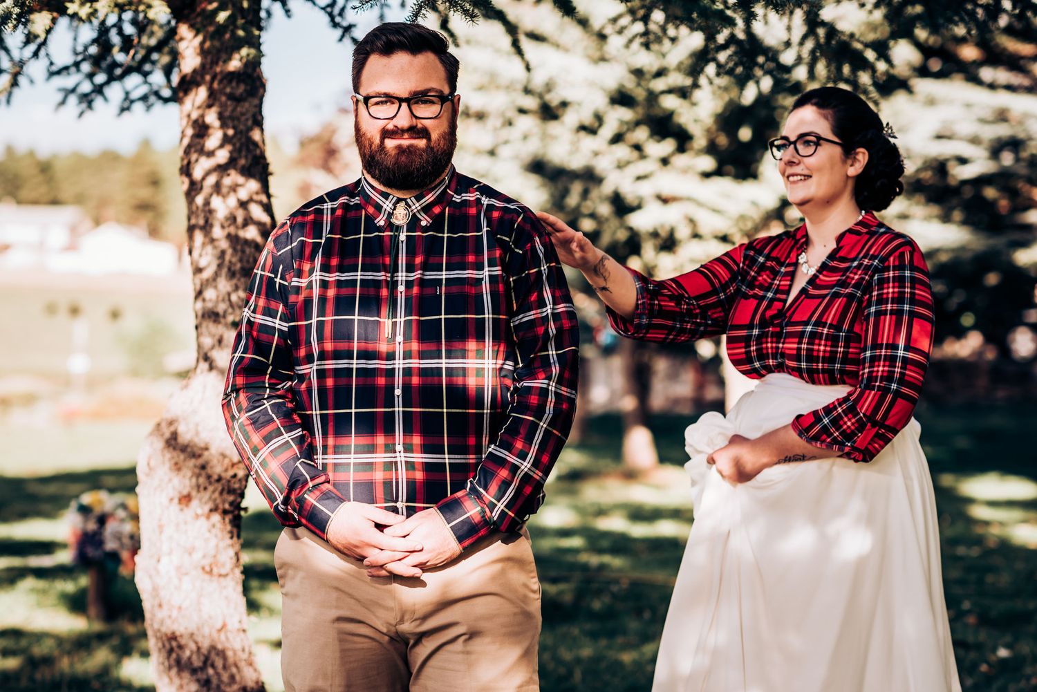 Couple in matching plaid shirts pose outdoors, one wearing a white skirt.