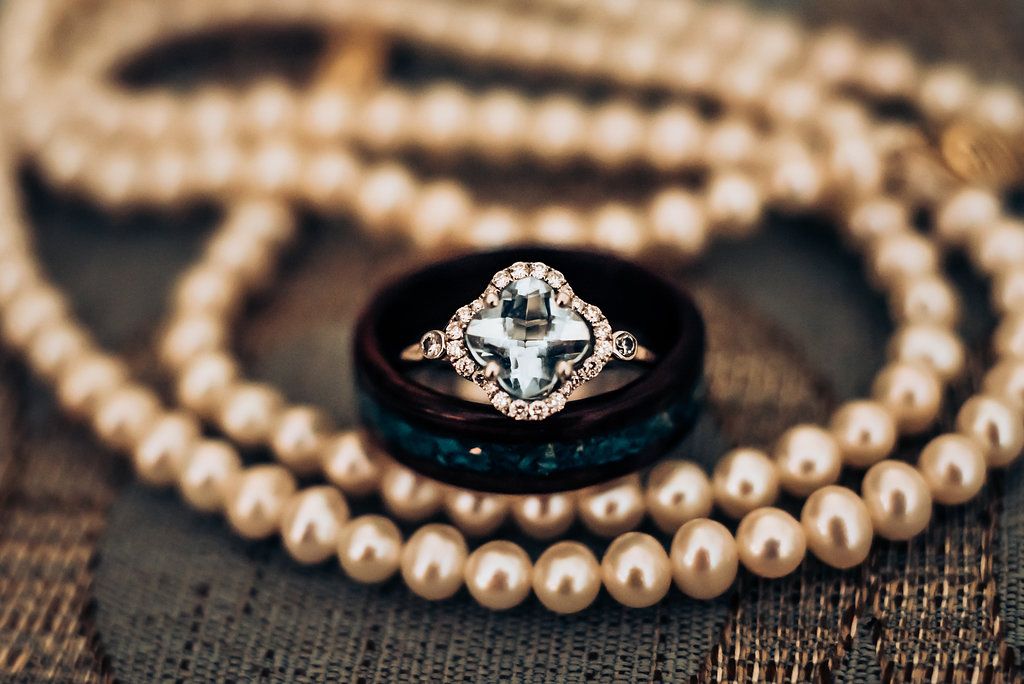 An elegant engagement ring surrounded by a pearl necklace on dark textured surface.
