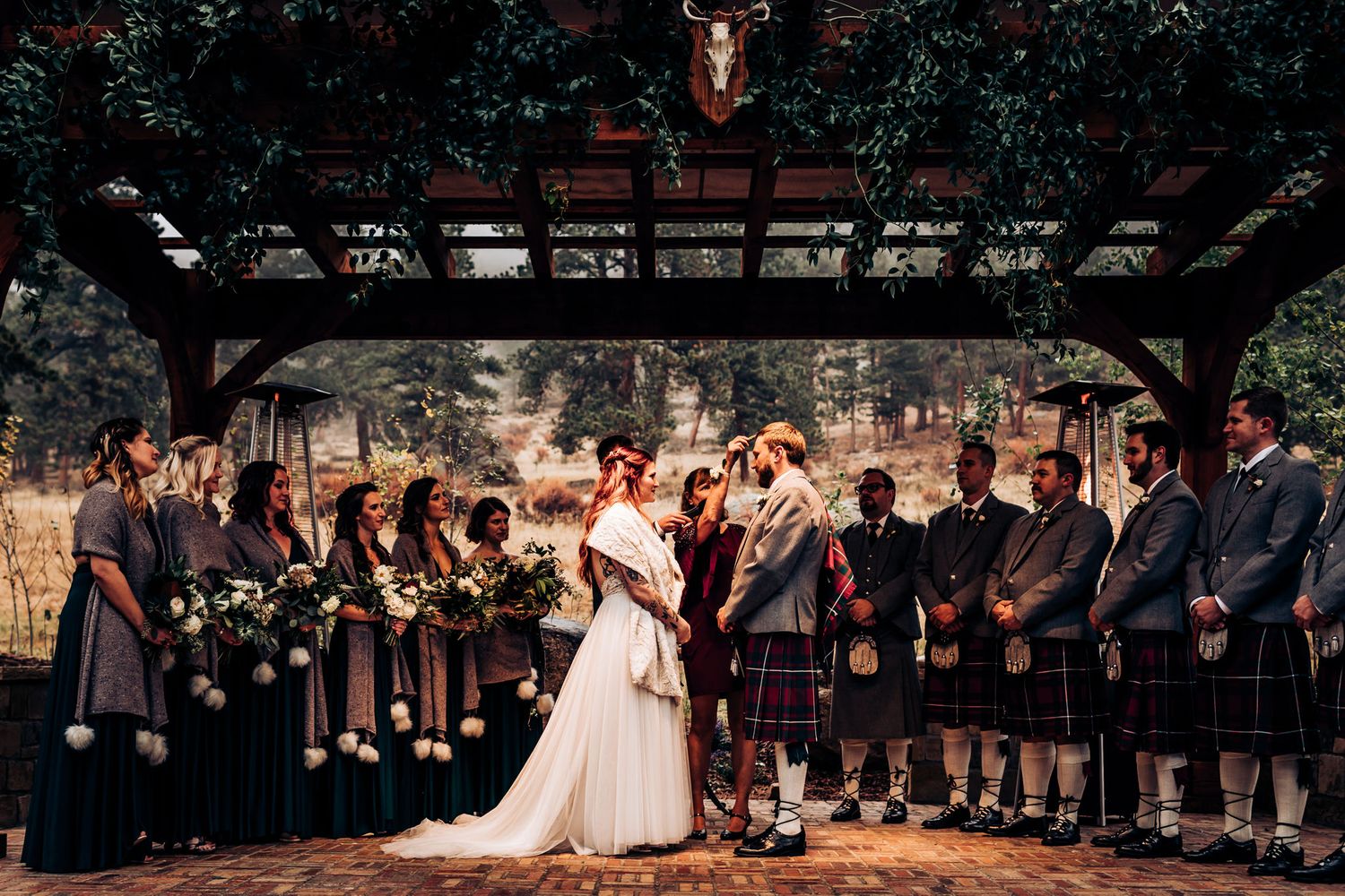 Bride in white dress stands with wedding party wearing kilts and formal attire under wooden pergola.
