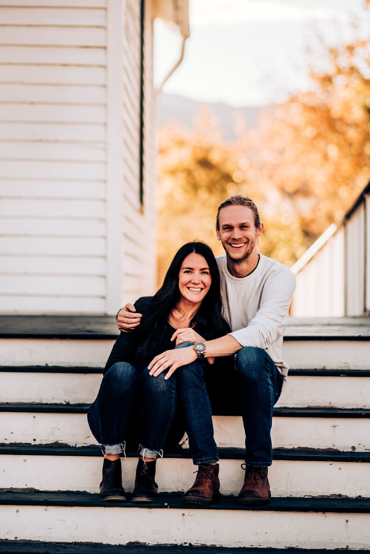 A couple embraces while sitting on white porch steps of a house in autumn.