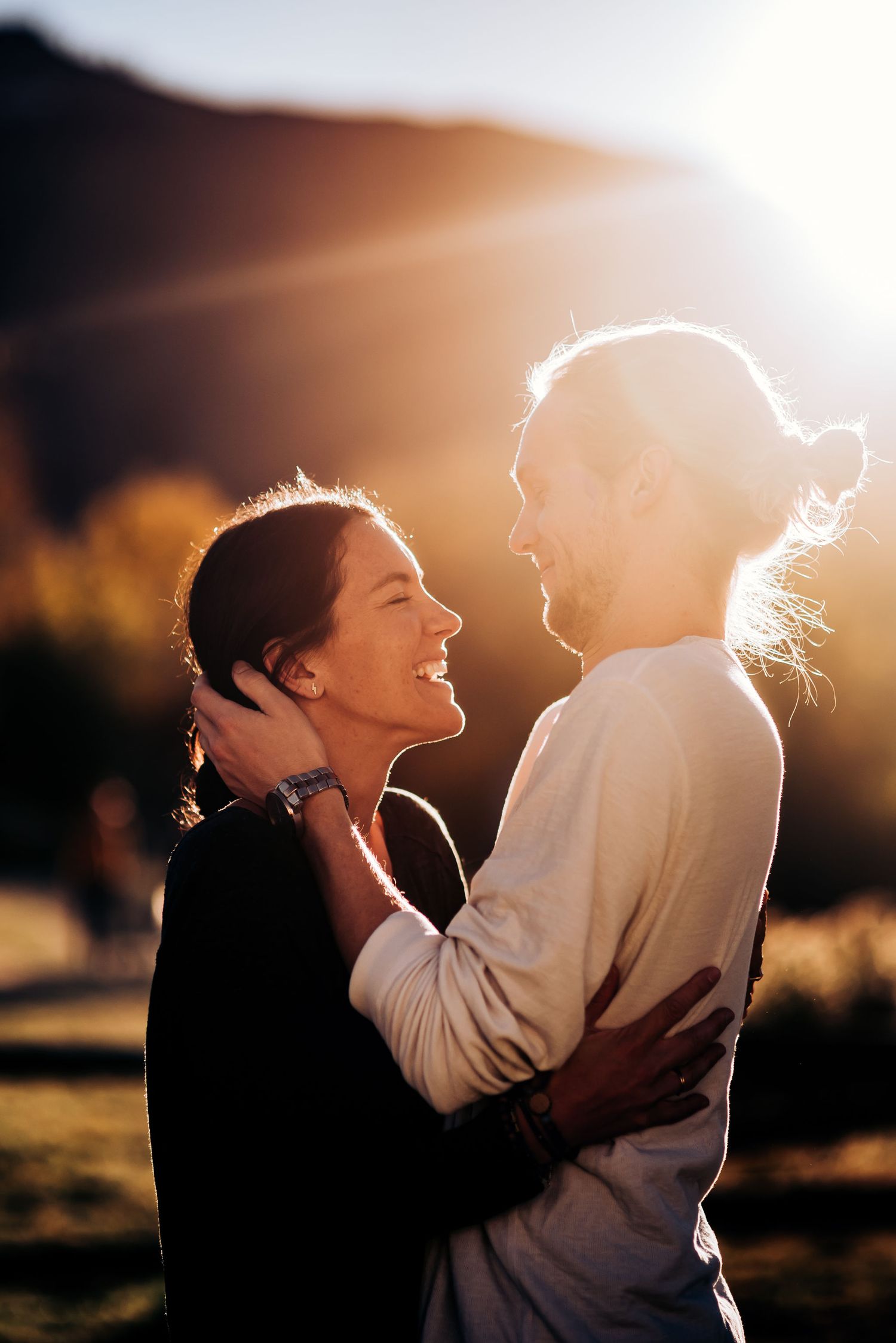 A joyful couple laughs together in warm sunlight with mountains visible behind them.